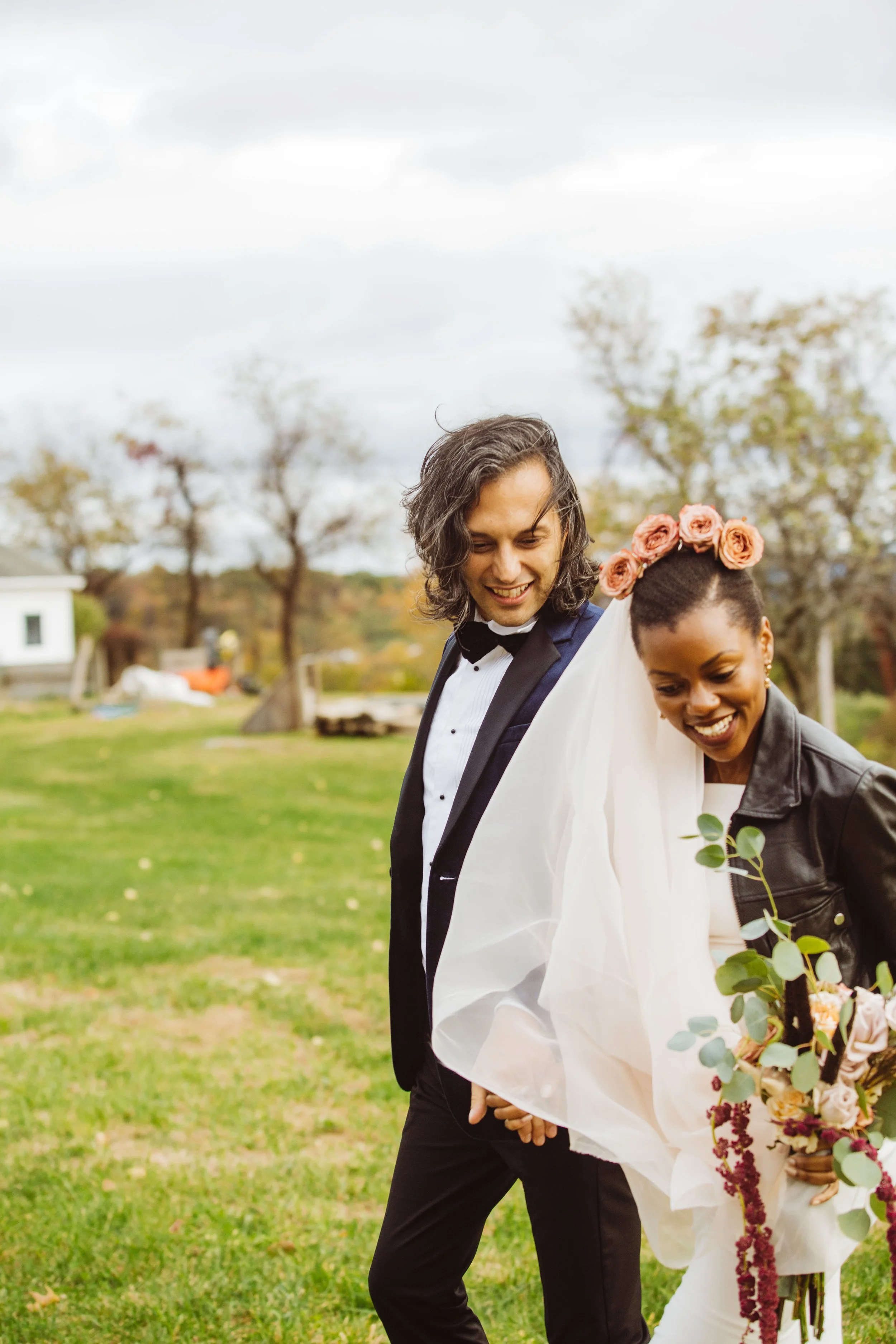 A smiling couple, dressed in wedding attire, walking outdoors on a cloudy day, with trees and a small building in the background. The woman has flowers in her hair and is holding a bouquet, while the man is wearing a tuxedo.