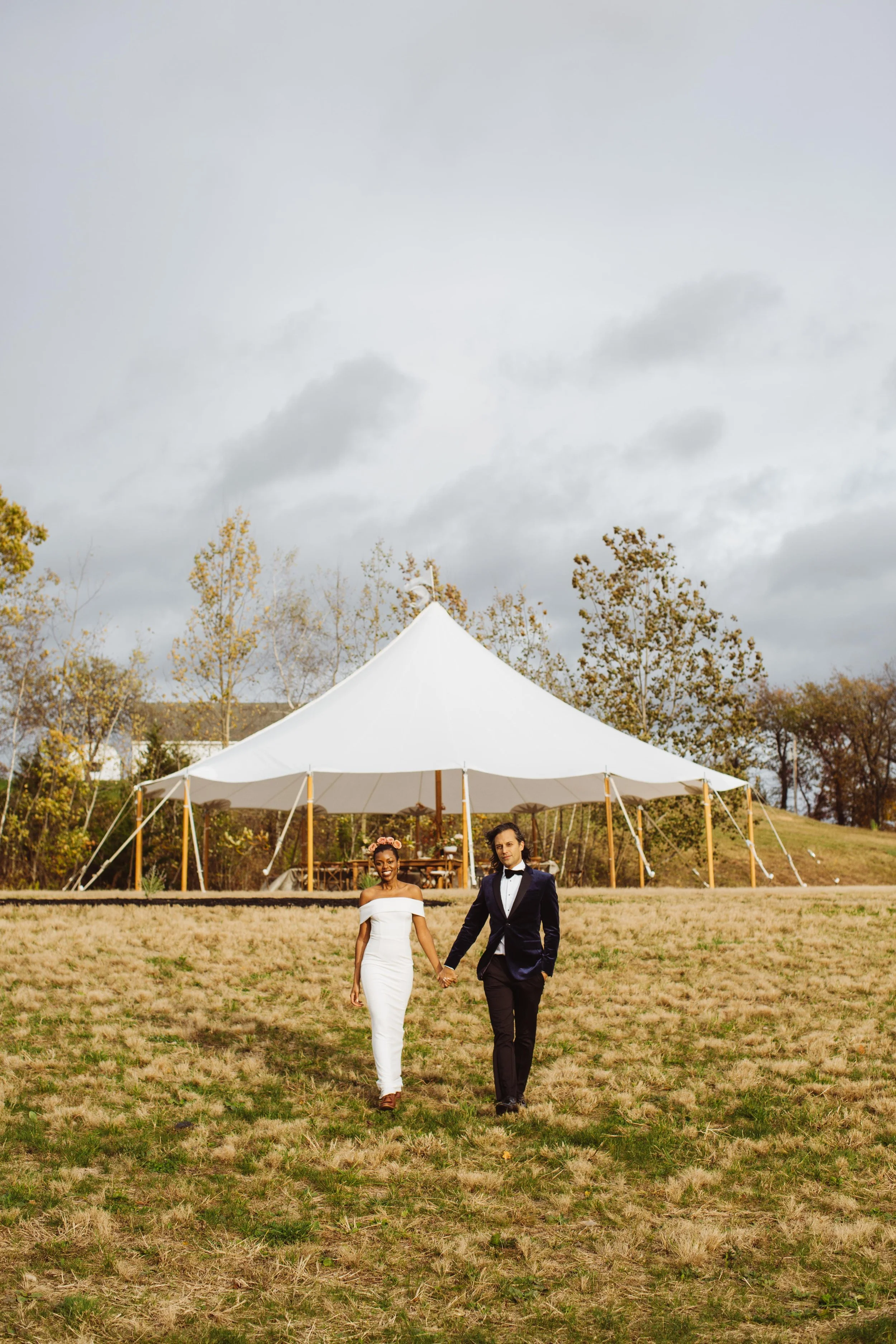 A couple walks hand-in-hand across a grassy field in front of a white wedding tent, with trees and cloudy sky in the background.