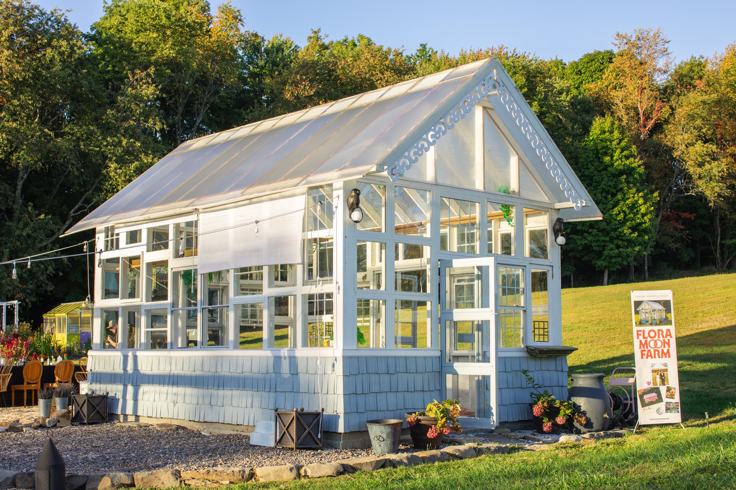 A white greenhouse with a gable roof, glass windows, and decorative trim located in a garden with trees and grass, with a sign that reads 'Flora Moon Farm' next to it.