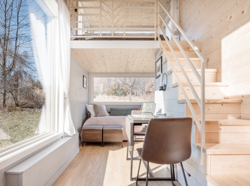 Interior view of a cozy tiny house with a large window, a small dining table with a brown chair, a pink sofa, and a lofted area with railing accessible by a staircase.