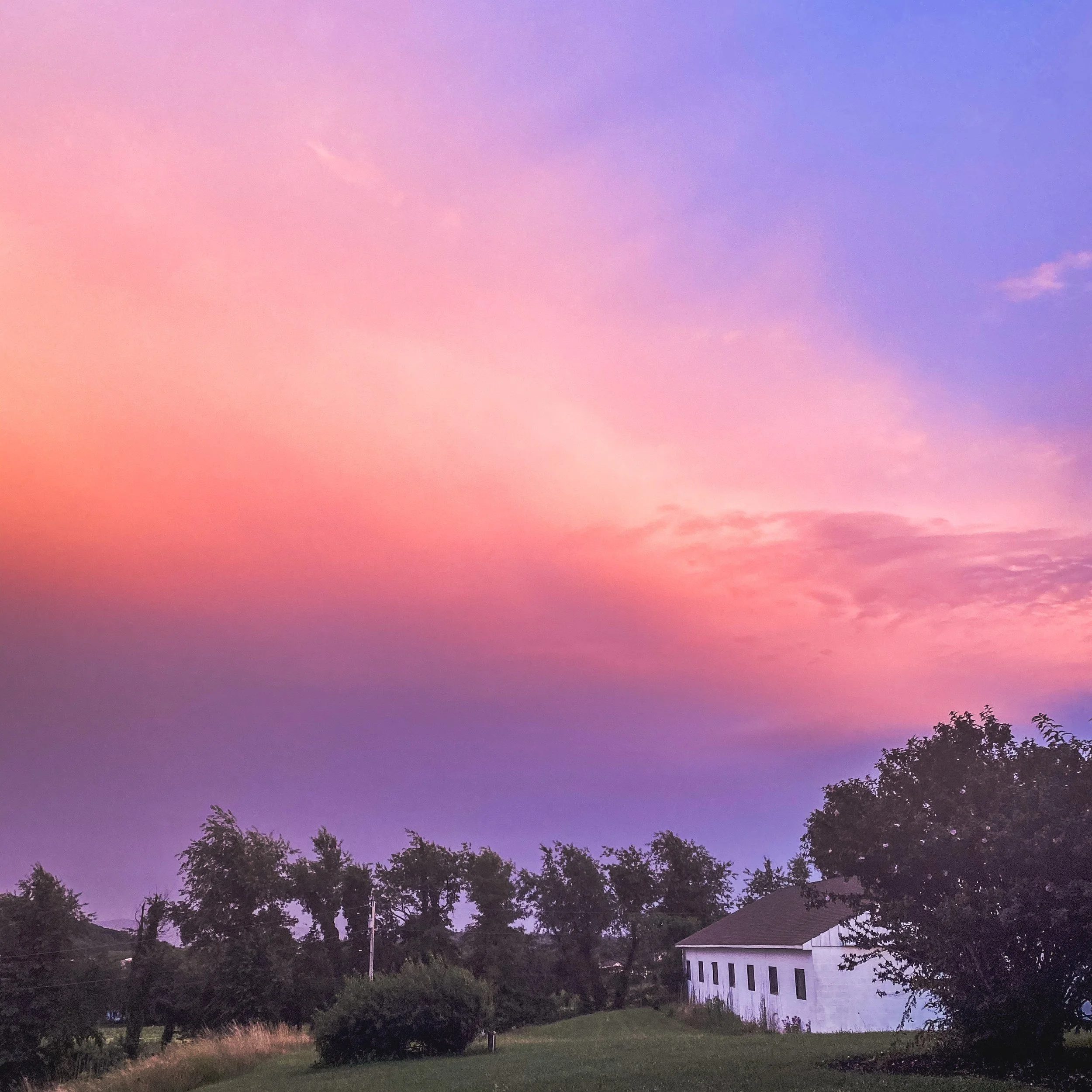 A colorful sky with pink, purple, and blue clouds over a grassy landscape with trees and a white building.