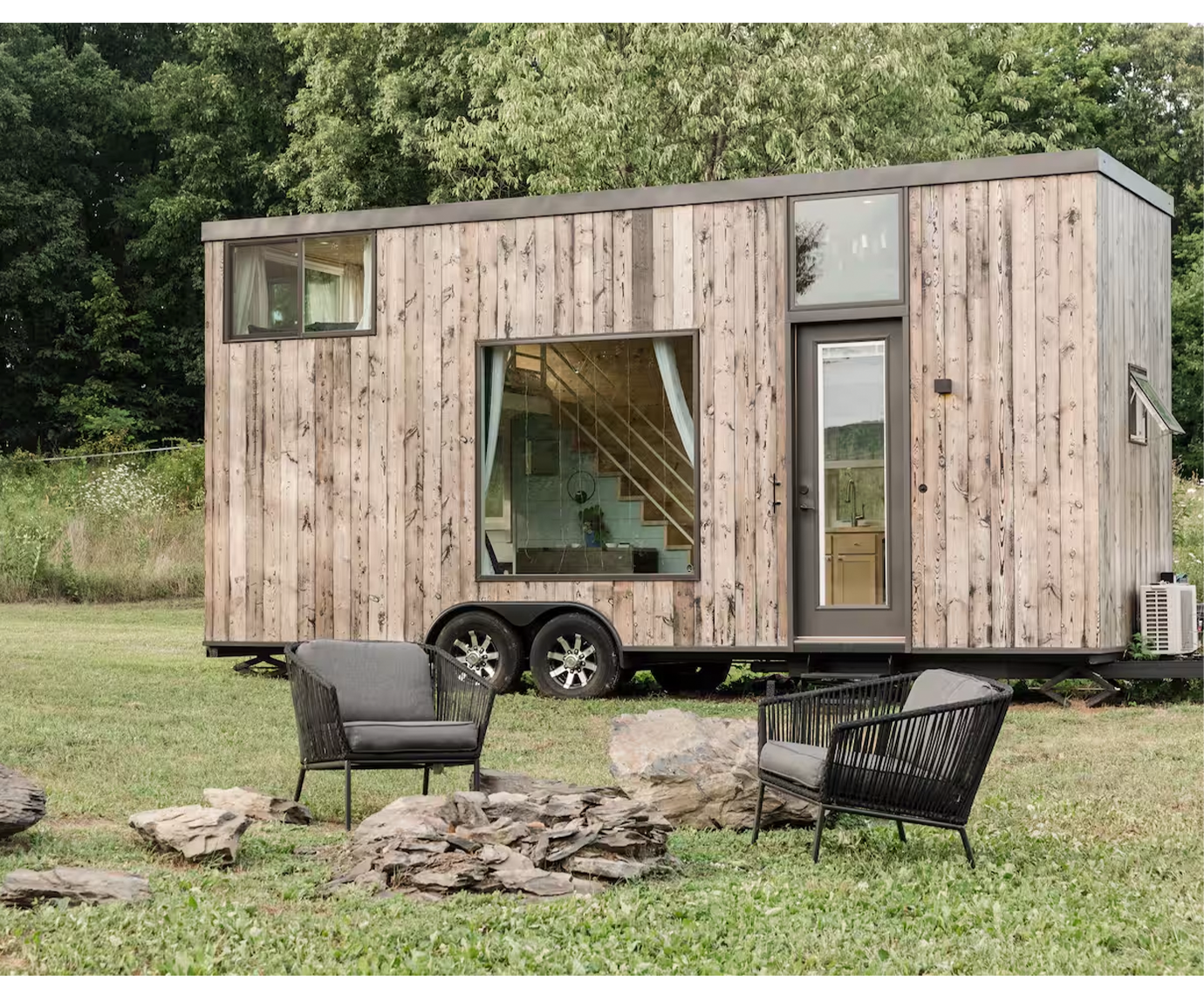 A small, modern wooden tiny house on wheels with large front window, side door, and small windows, situated in a grassy yard with outdoor seating and a fire pit.