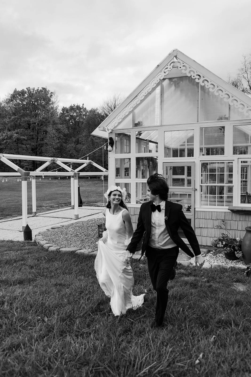 A newlywed couple holding hands and running outside near a glass greenhouse.