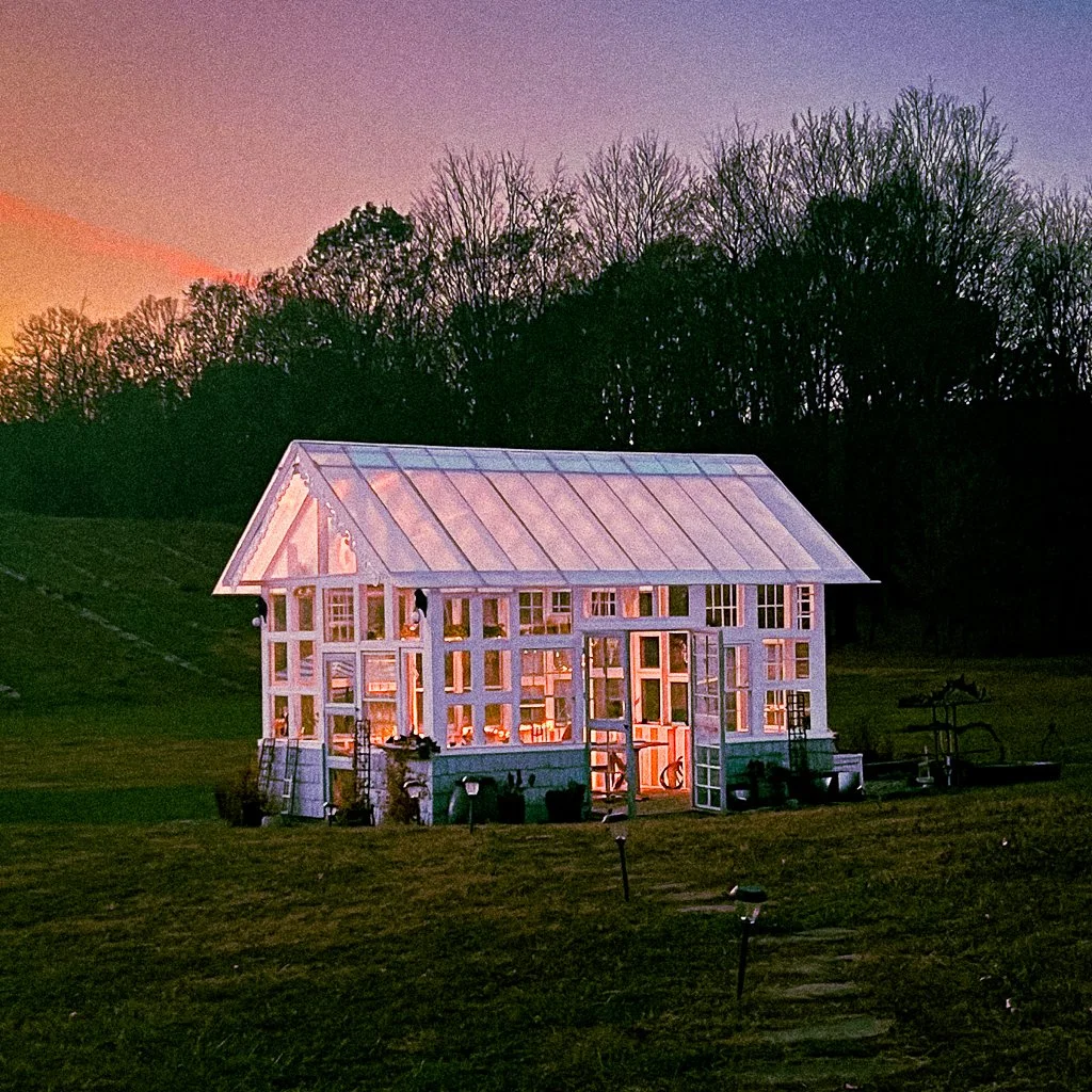 A modern glasshouse or greenhouse with a balcony, illuminated from within, set on a grassy field during sunset with trees in the background.