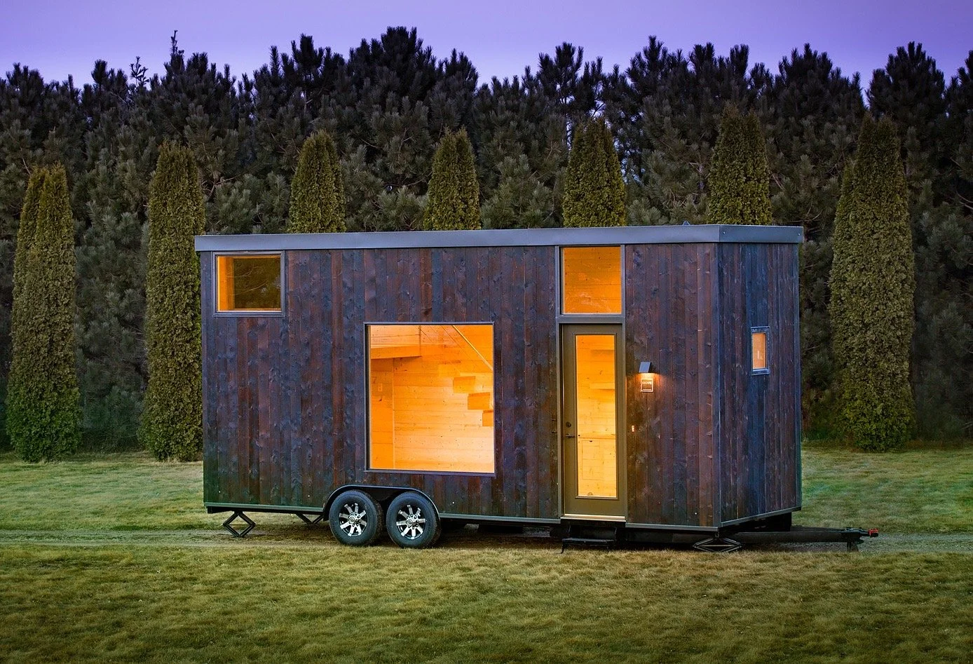 Tiny modern house trailer with dark wood exterior and illuminated interior, situated on grass with tall trees in the background at dusk.