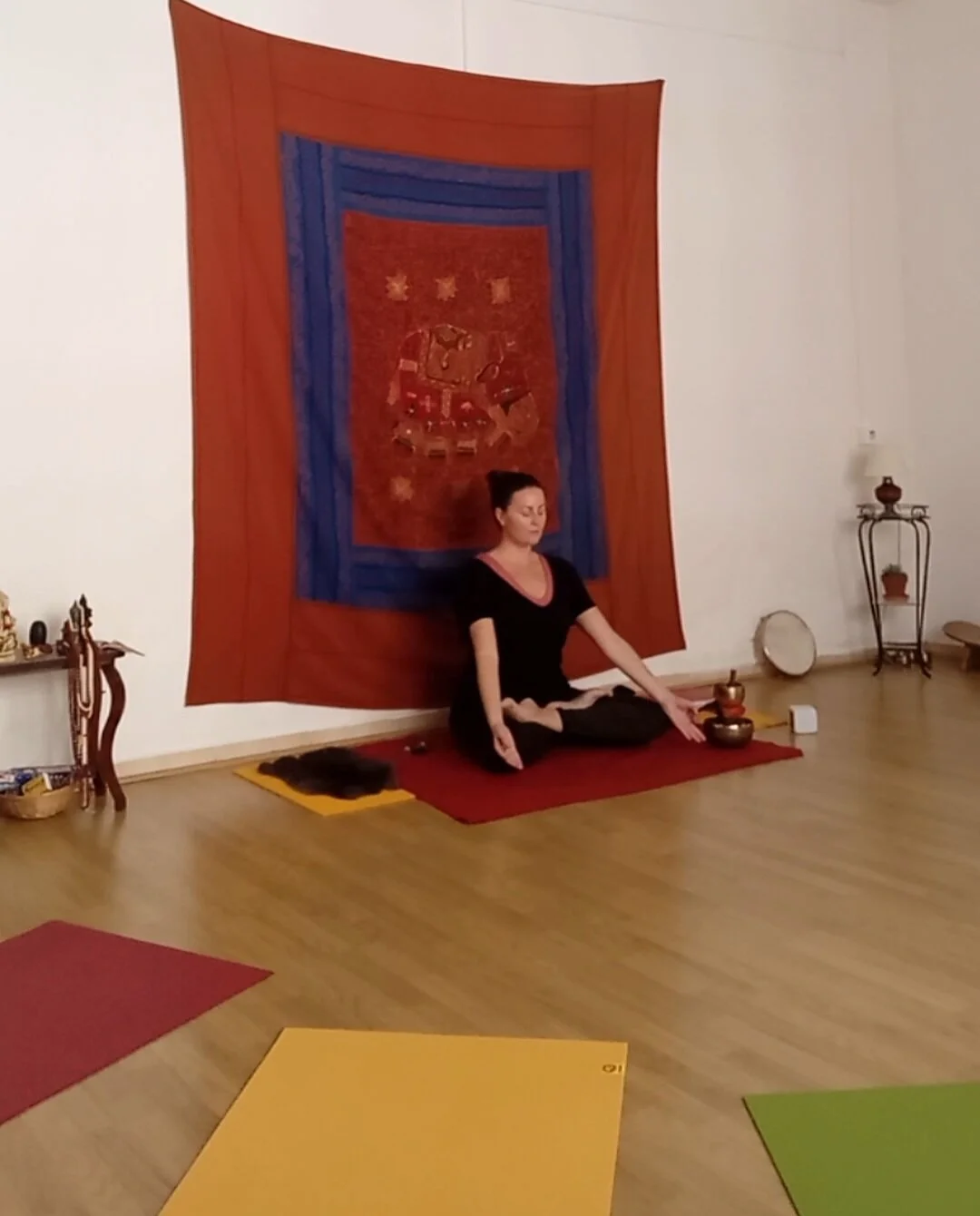 Laure pratiquant la méditation ou le yoga en position assise dans une pièce avec un tapis rouge, devant un grand tapis mural coloré, entourée de petits accessoires et décorations zen.