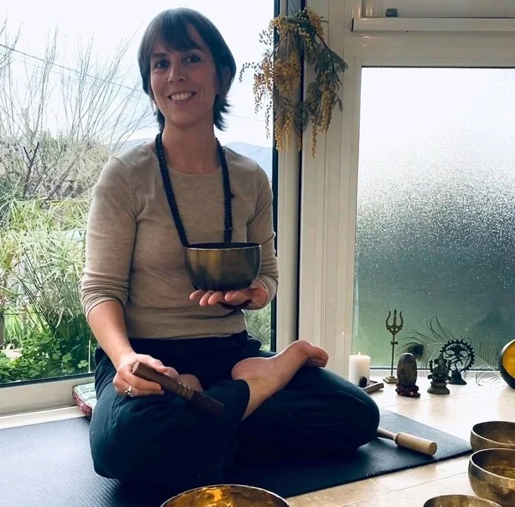 Agathe, souriante, assise en position de lotus sur un tapis de yoga, tenant un bol en métal dans une main et un rituel en bois dans l'autre, dans une pièce lumineuse avec des objets religieux ou spirituels et un jardin visible par la fenêtre.