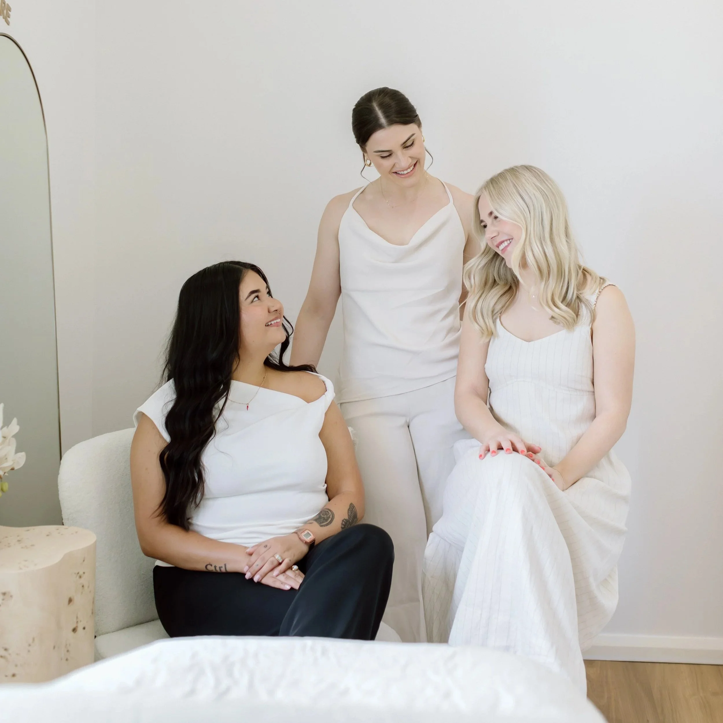 Three women in white outfits smiling and interacting indoors, with two seated and one standing, in a bright, minimalistic treatment room.