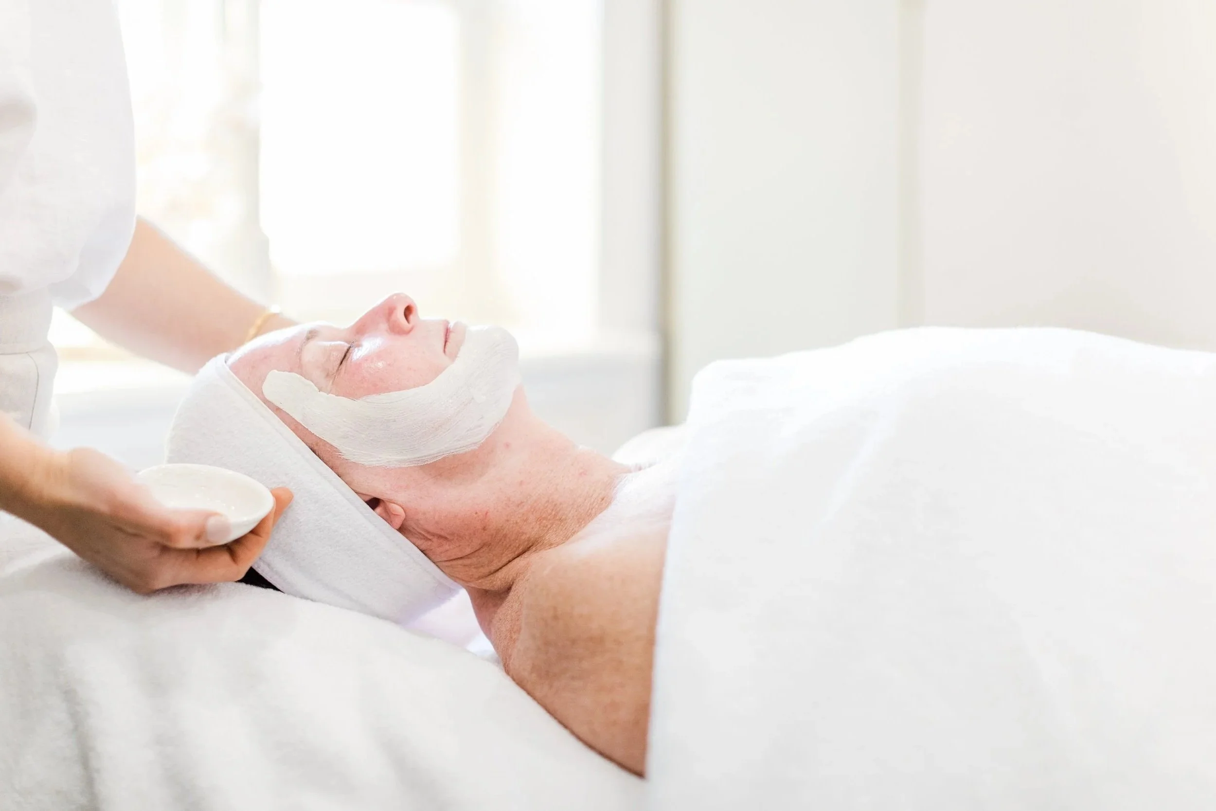 A woman receiving a facial treatment at a spa, with a white facial mask being applied and a towel wrapped around her head, lying on a treatment table.