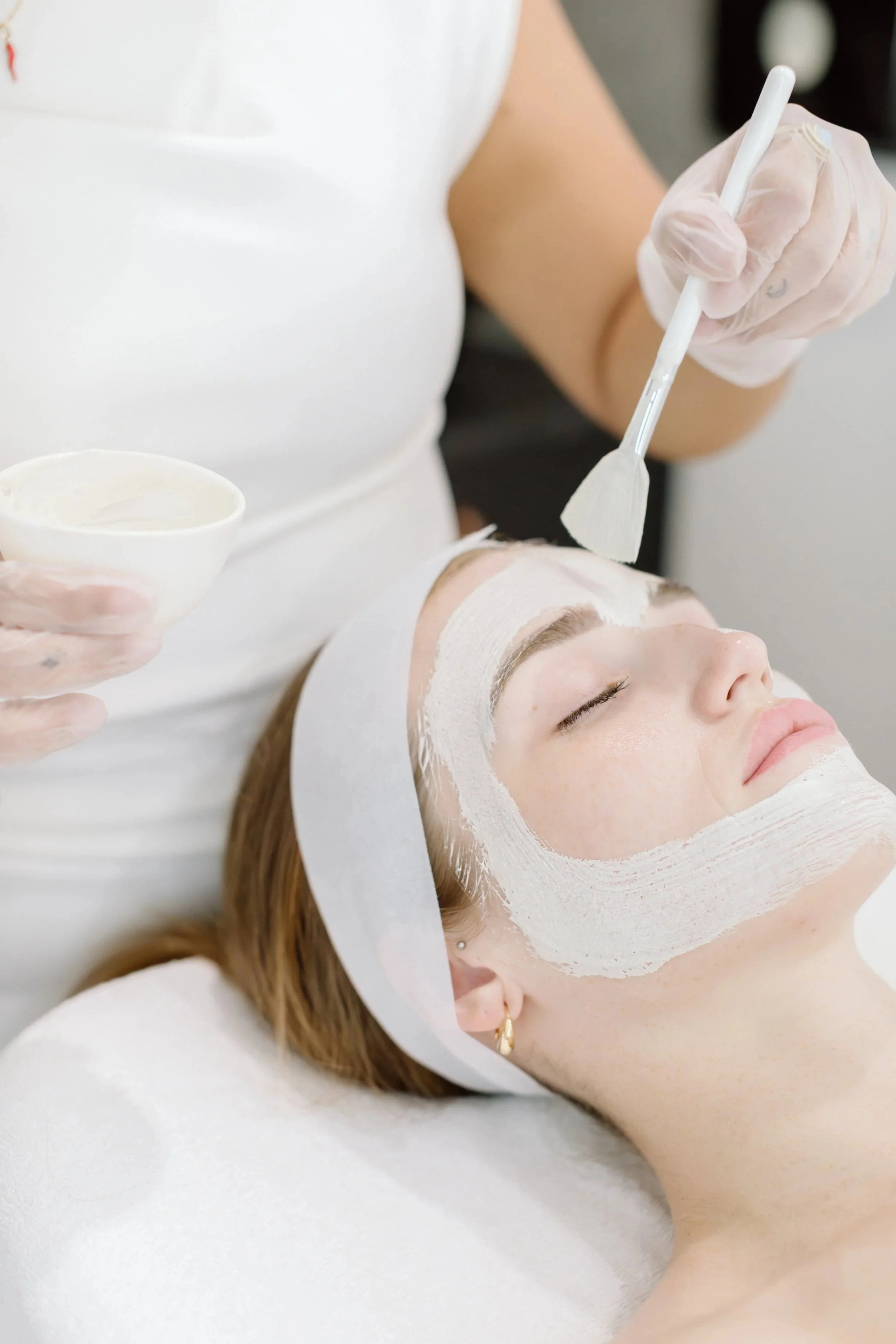 A woman receiving a facial treatment, lying with a facial mask being applied on her face, wearing a headband, in a spa or clinic environment.