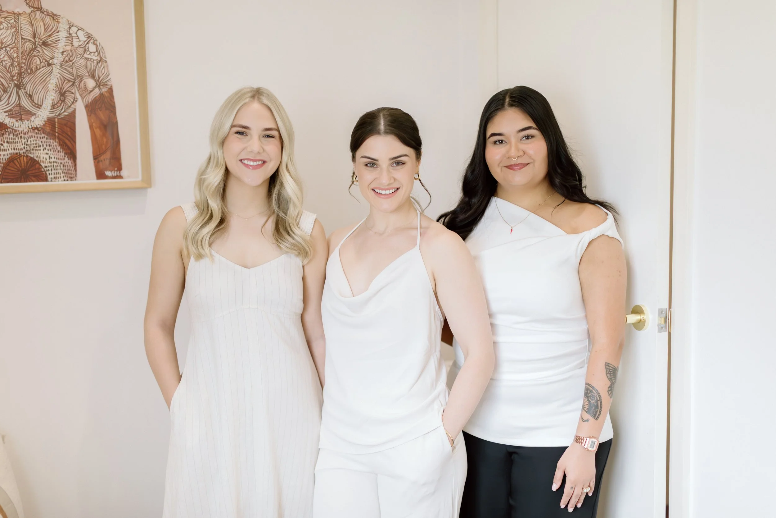 Three women standing together indoors, smiling, wearing white tops and dark bottoms, with a framed abstract artwork hanging on the wall behind them.