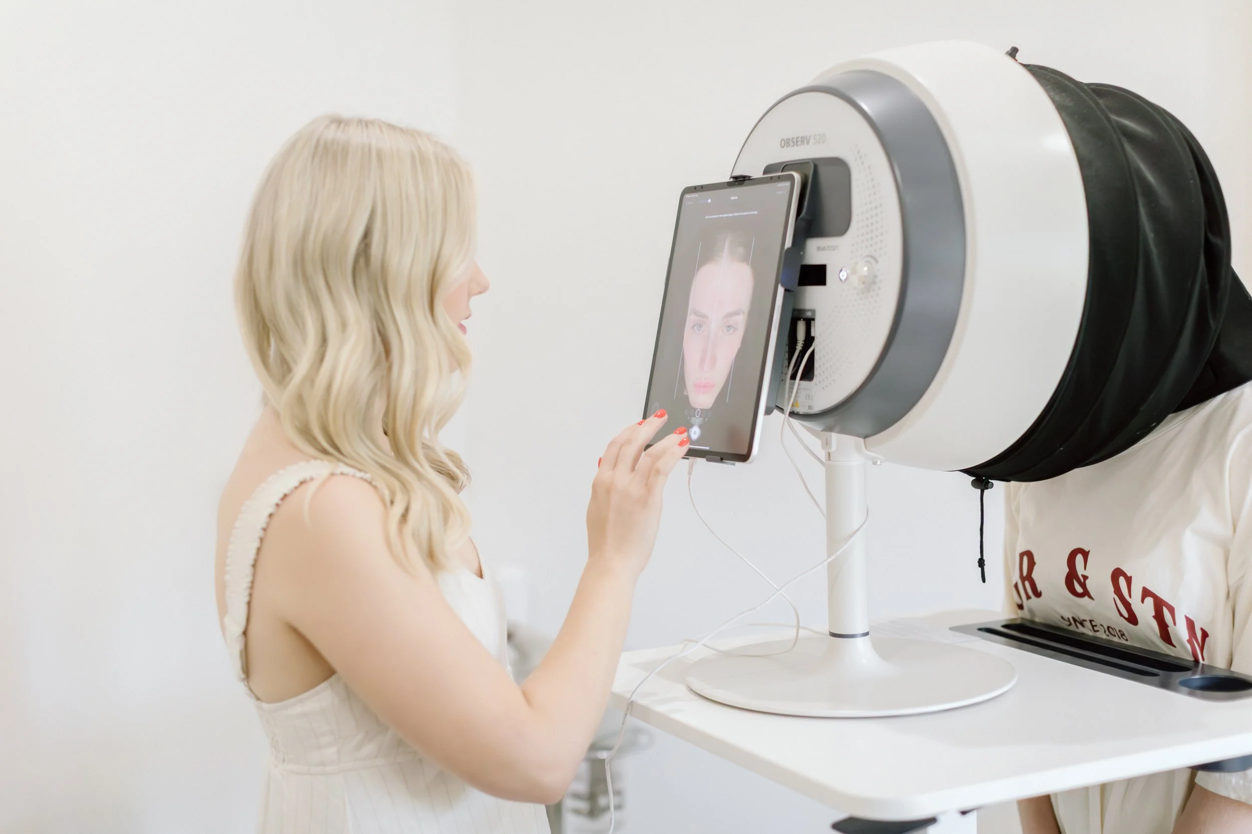 A woman with blonde hair looking at a tablet that shows a face inside a skin scanning machine.