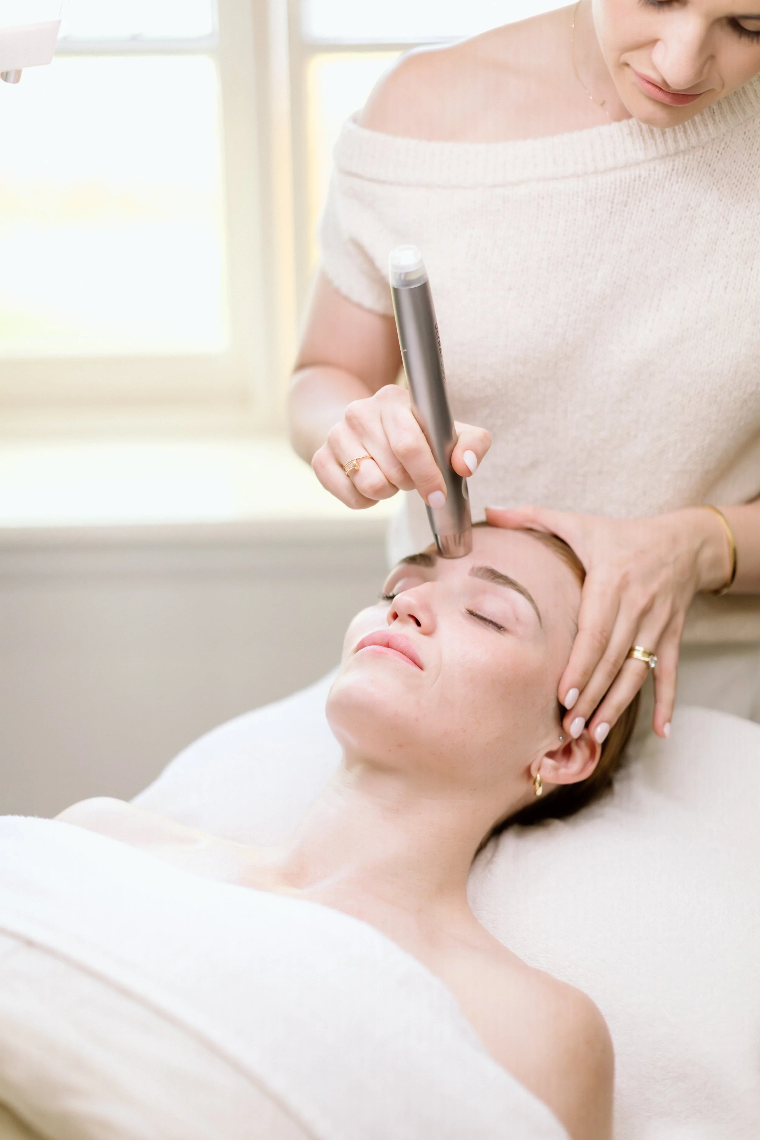 A woman receiving a facial treatment with a handheld tool applied to her forehead while lying down with her eyes closed.