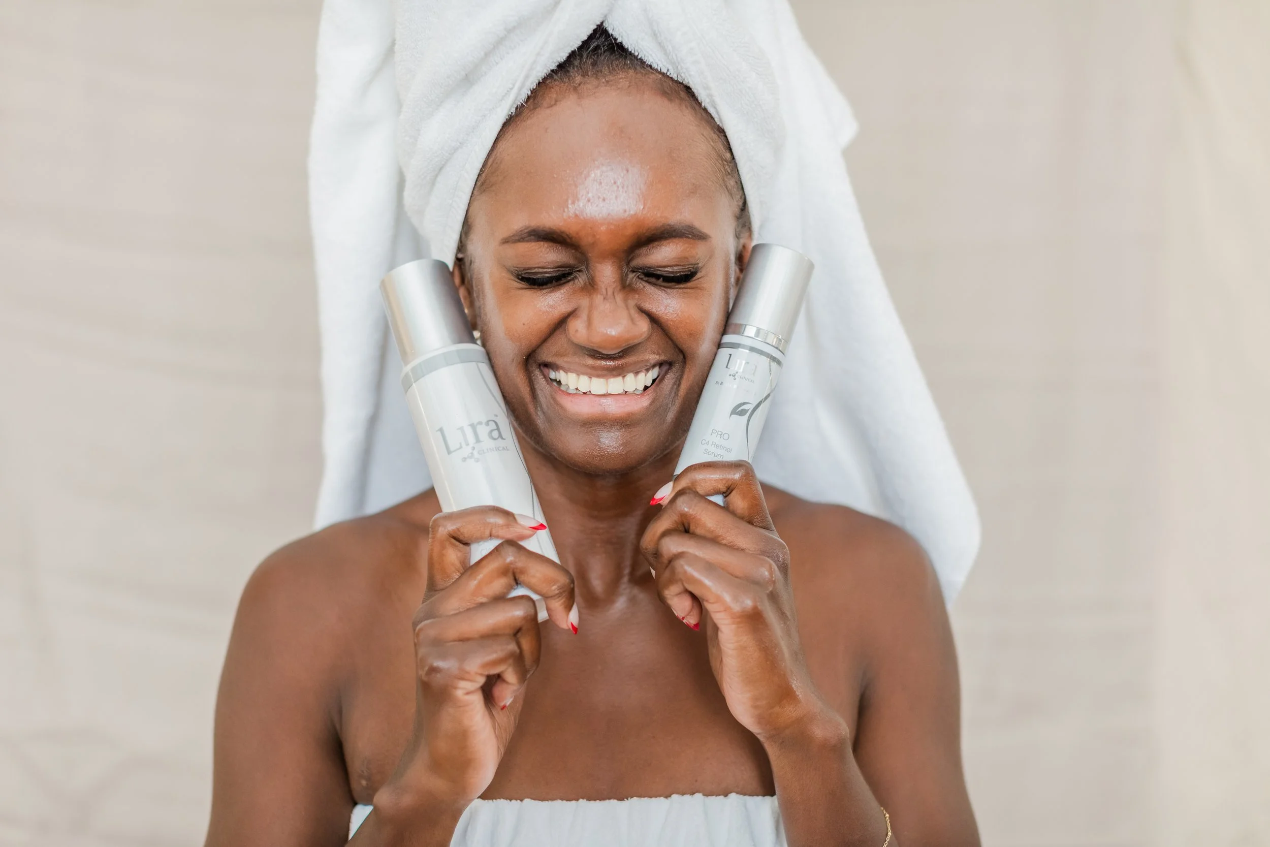 Smiling woman with a towel wrapped around her hair, holding two skincare serum bottles, one in each hand, in a bright room.