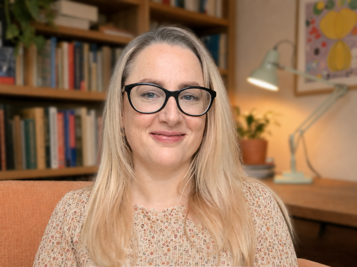 A woman with blonde hair and black glasses smiling at the camera, seated in a cozy, well-lit room with bookshelves, a desk lamp, and plants in the background.
