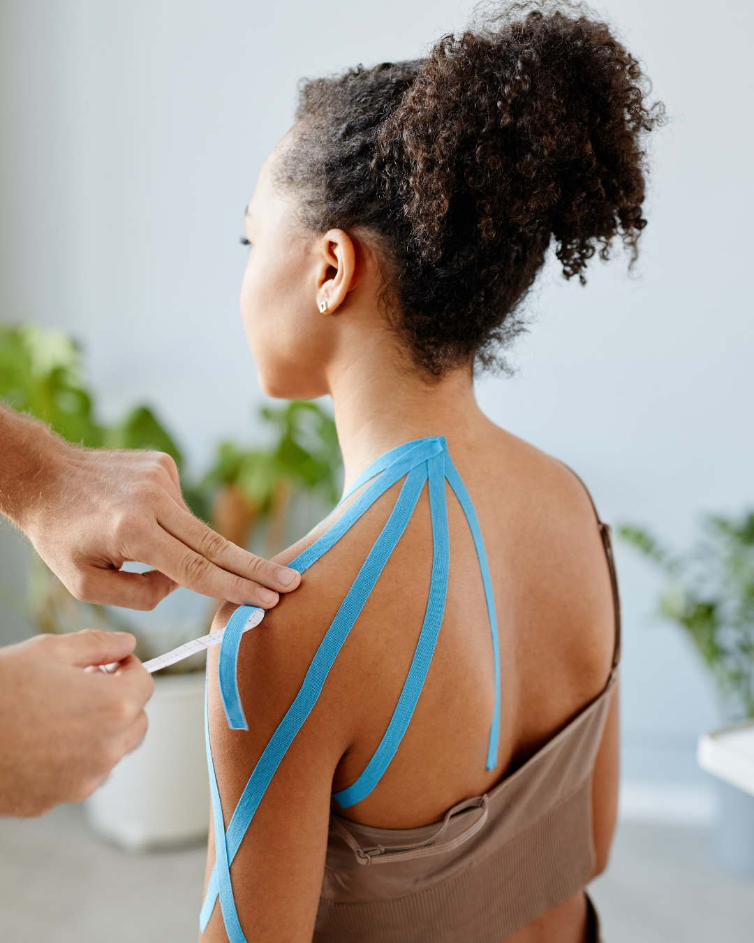 A woman having her shoulder measured with a blue measuring tape by a person, in a setting with green plants in the background.