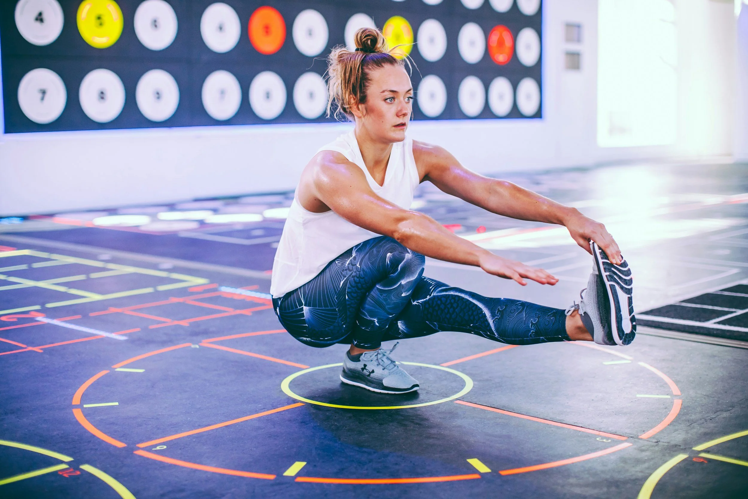 A woman in workout clothes is performing a stretching exercise in a gym with colorful markings on the floor. She is balancing on one foot while extending her other leg and reaching for her foot with her hand. The gym has a sports-themed background wall.