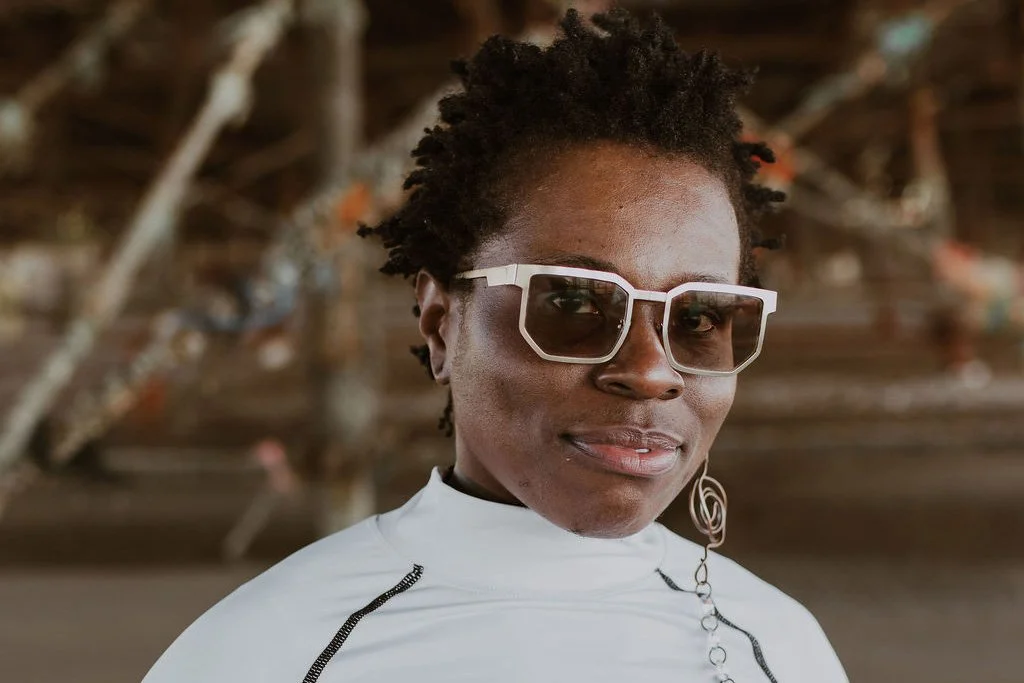 A black queer person dressed in white with white rimmed glasses smiling