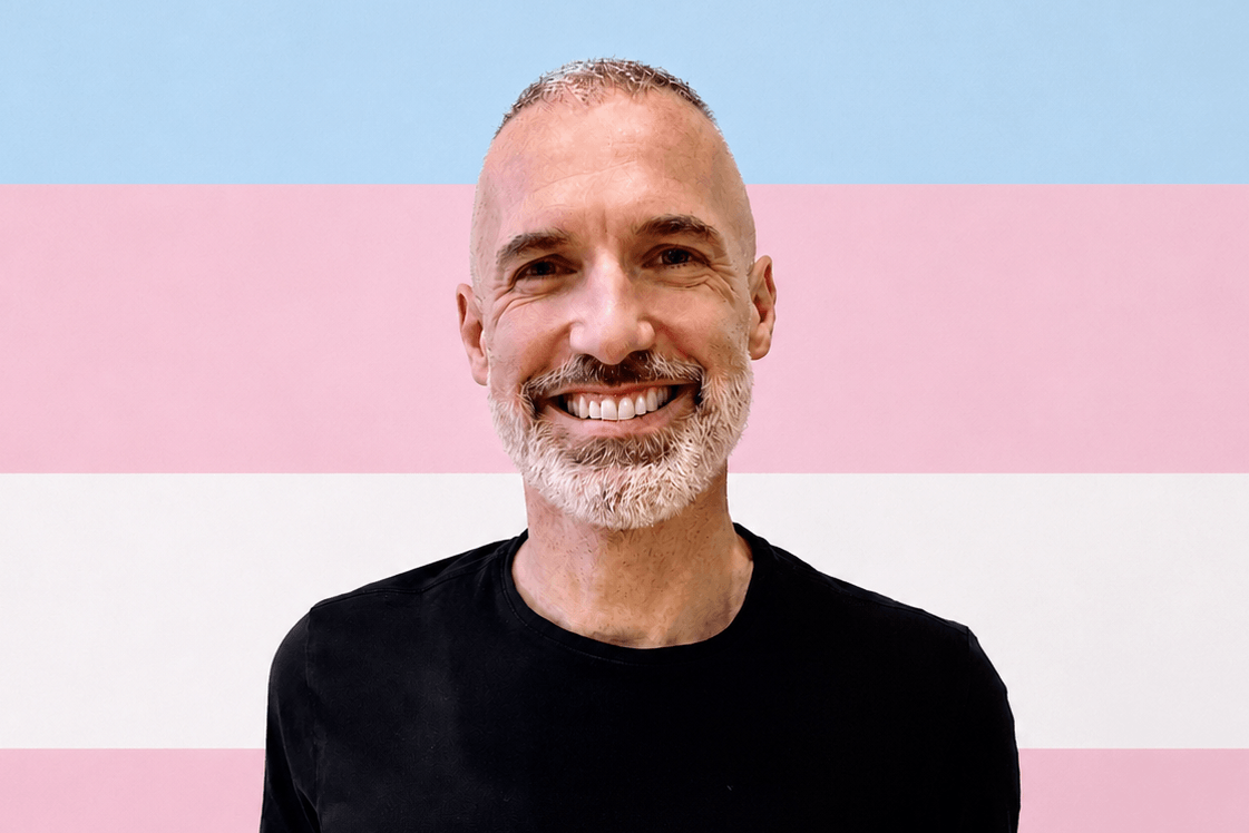 A person with a short white beard smiling in front of a trans flag.