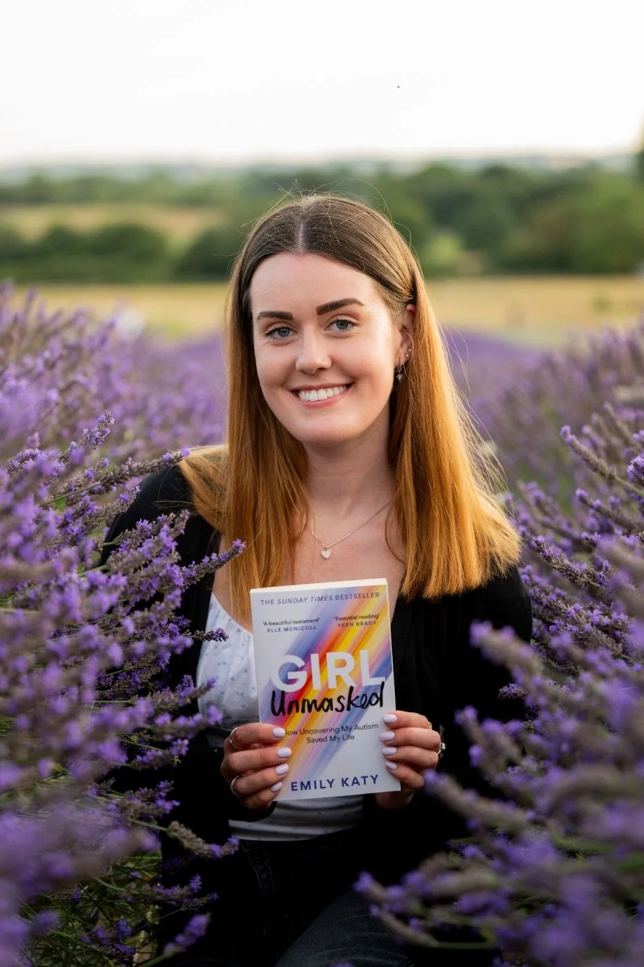 Emily Katy smiling in a lavender field, holding her book Girl Unmasked
