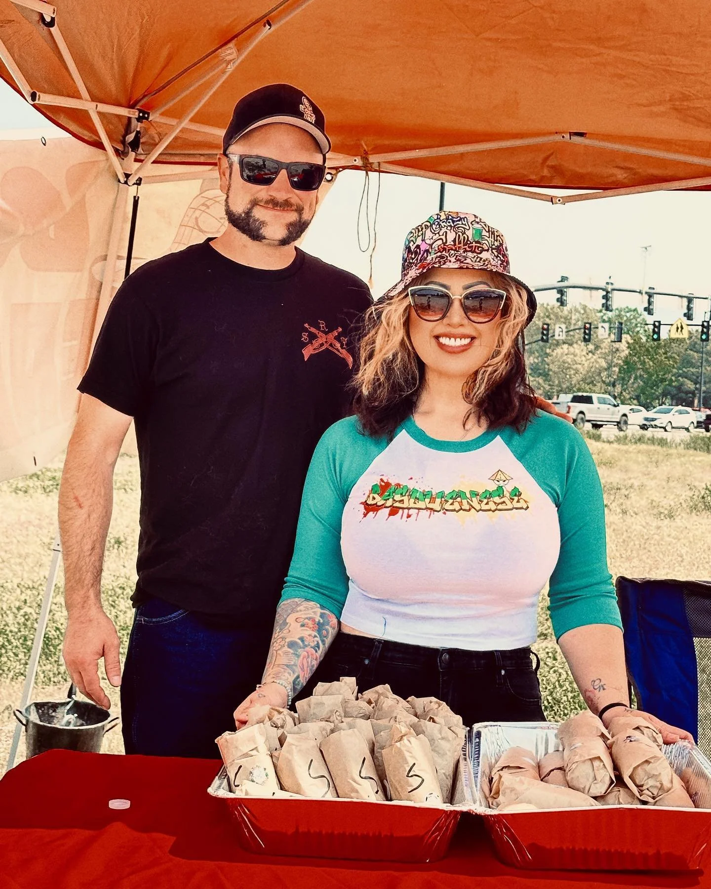 A man and woman standing behind a table with food wrapped in paper at an outdoor event under a beige canopy, with traffic and trees in the background. The woman is smiling, wearing sunglasses, a colorful hat, a white and teal shirt, and has visible tattoos on her arms.