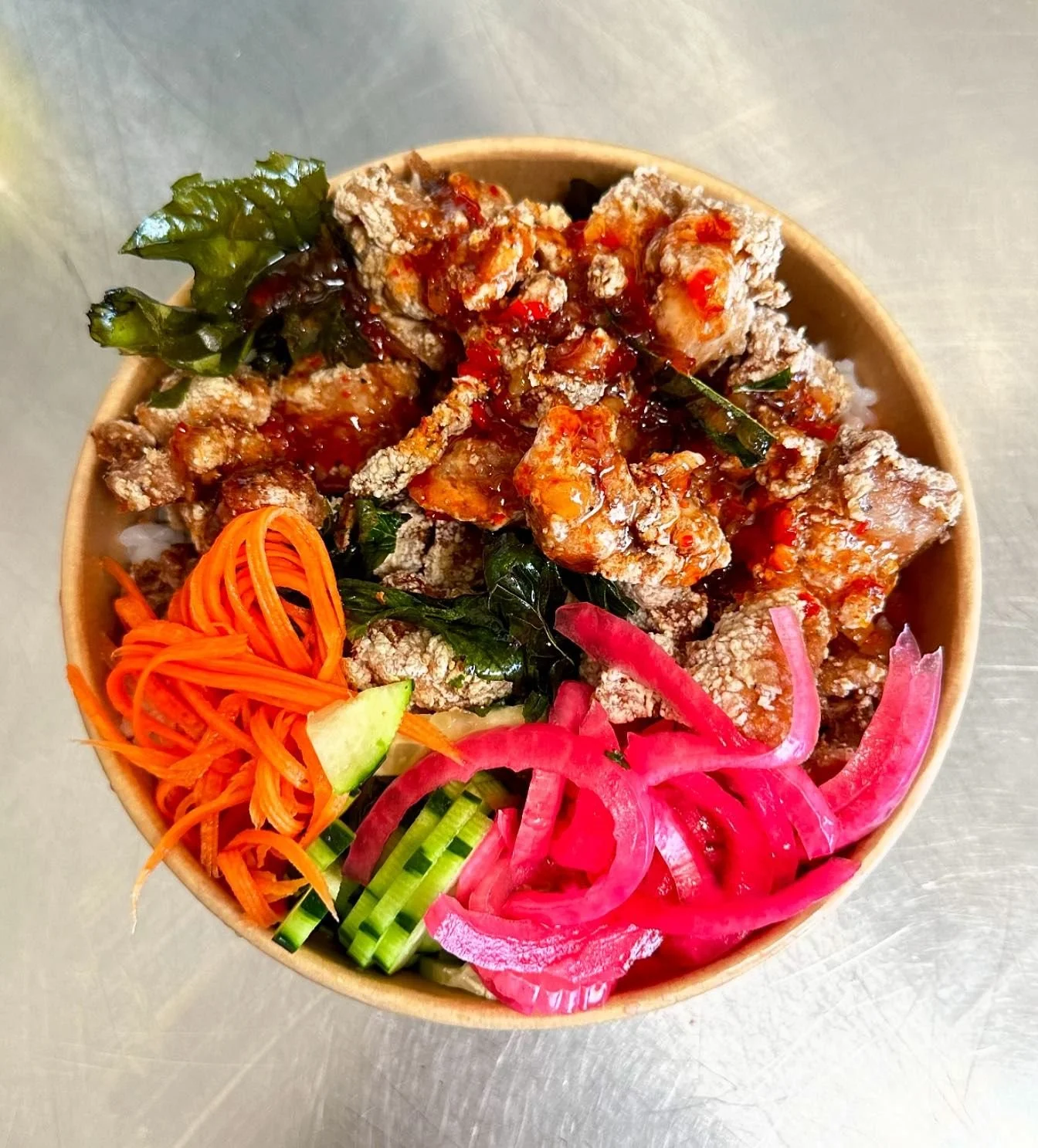 Bowl of rice topped with fried chicken, red sauce, and green herbs, accompanied by shredded carrots, sliced cucumber, and pickled red onions.
