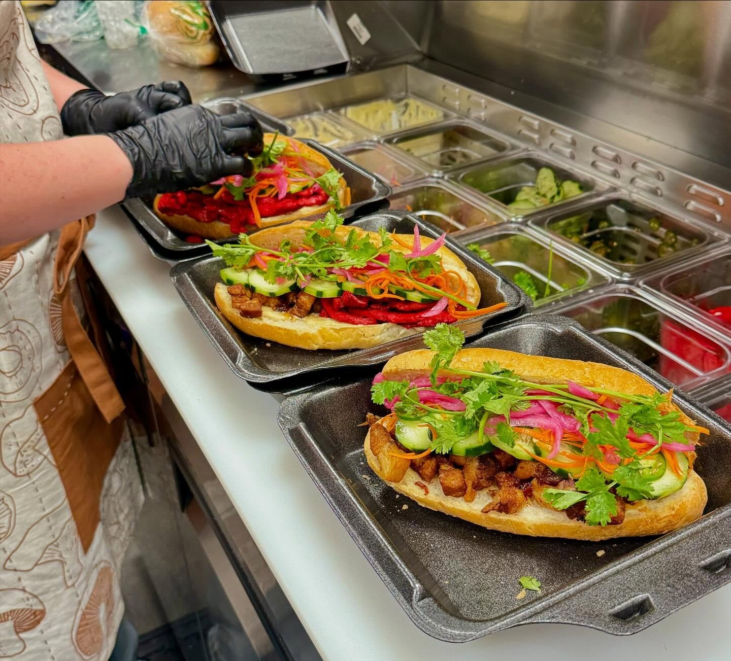 Three Vietnamese banh mi sandwiches with fresh vegetables and grilled meat, being prepared on a kitchen counter with various toppings in the background.