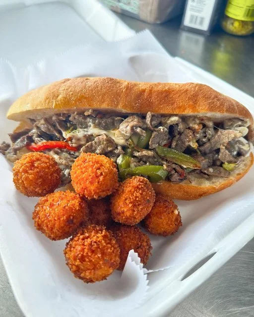A sandwich with sliced beef and green peppers in a bun, served with several fried, breaded cheese balls on a white tray.