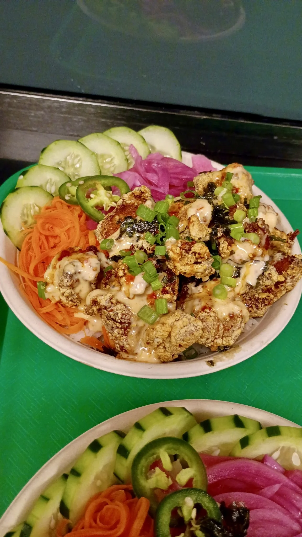 A bowl of fried chicken topped with green onions and sauce, served with sliced cucumbers, jalapeños, shredded carrots, and pickled red onions.