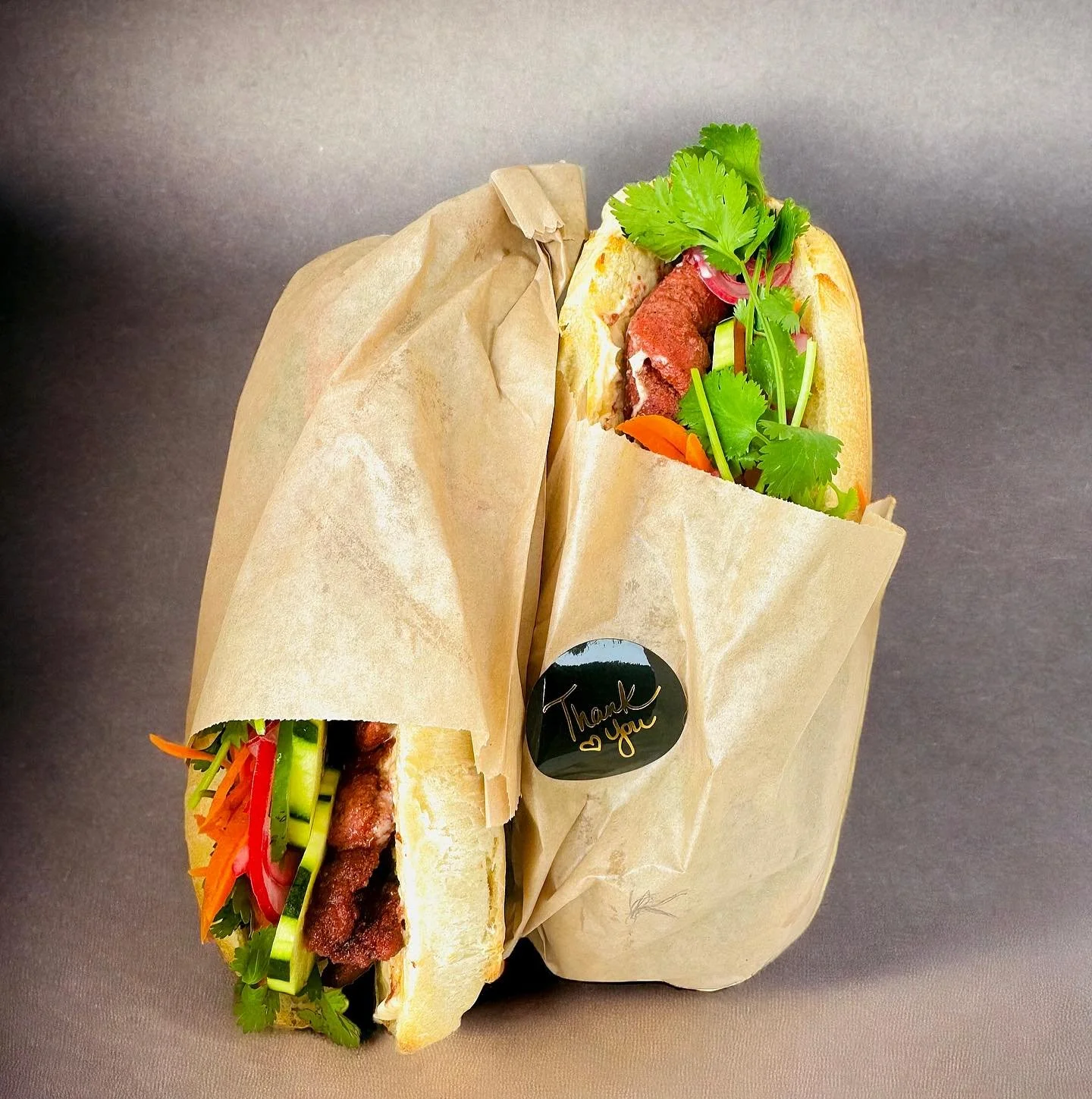 Two wrapped Vietnamese banh mi sandwiches filled with sliced cucumbers, carrots, red peppers, cilantro, and grilled meat, with a small black sticker that says "Thank you" and a heart symbol.