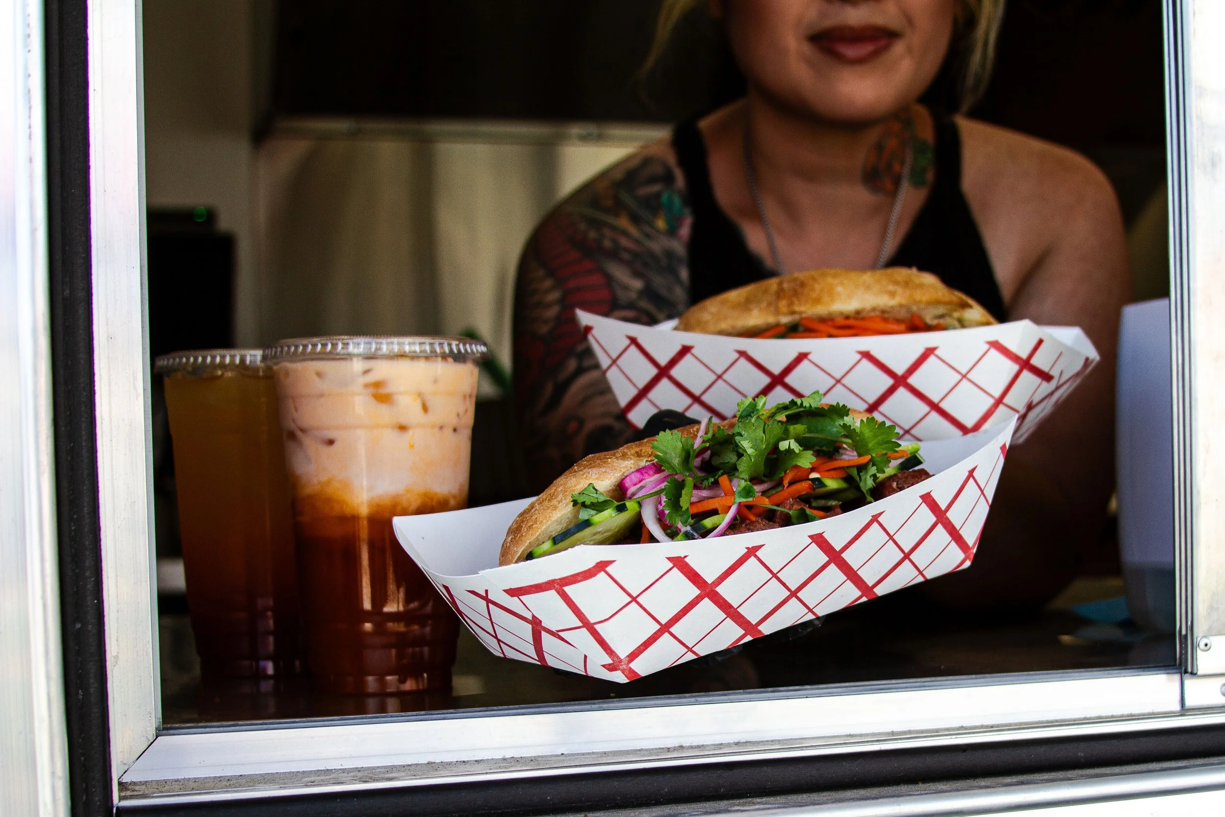 Woman with tattoos serving two sandwiches with fresh vegetables in white baskets, accompanied by a cup of iced tea or soda in a plastic cup, through a food truck window.