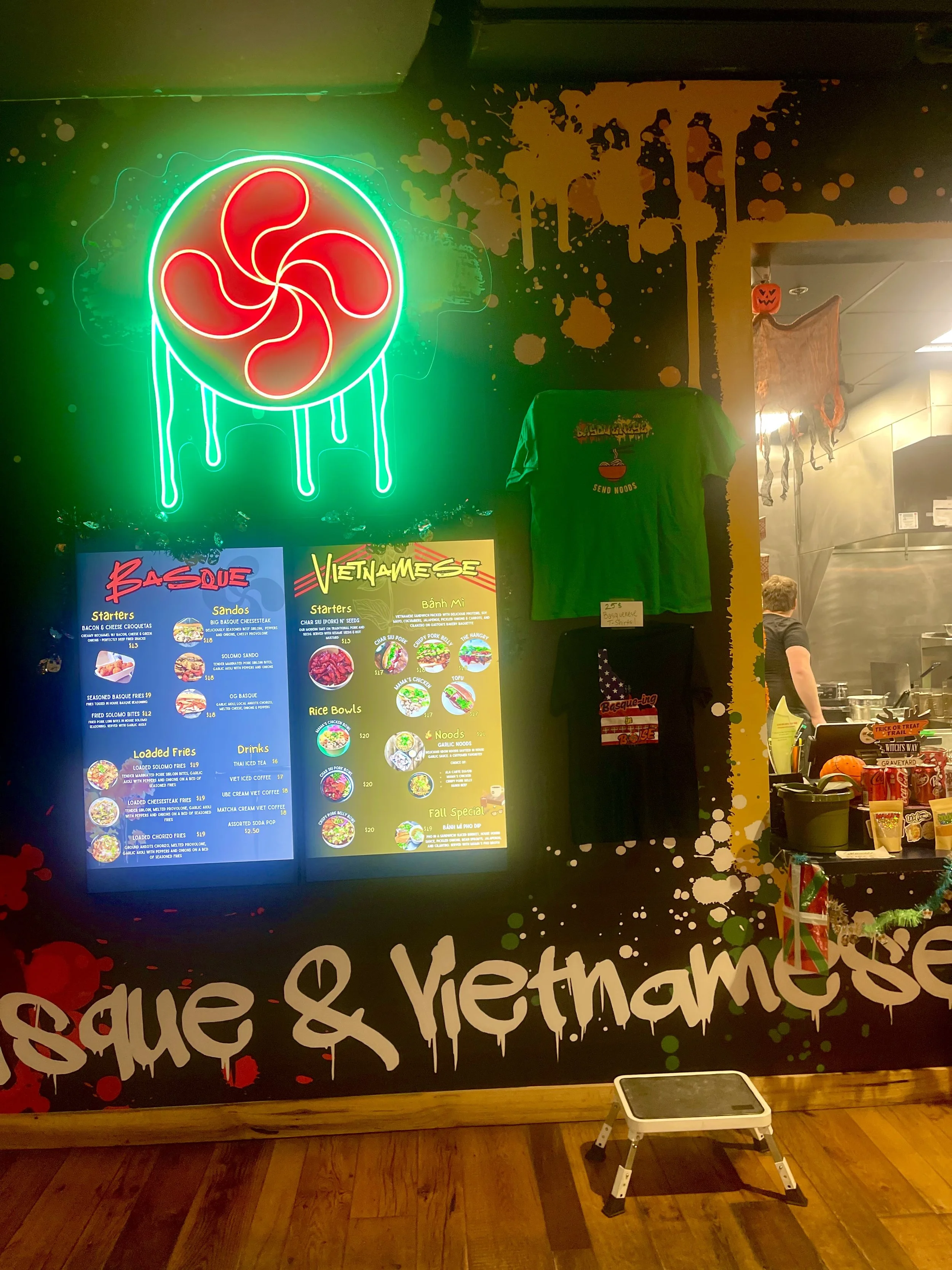 Neon sign of a red and green swirl flower with dripping effect, above a menu board and a green Vietnamese T-shirt hanging on the wall in a restaurant decorated for Halloween.