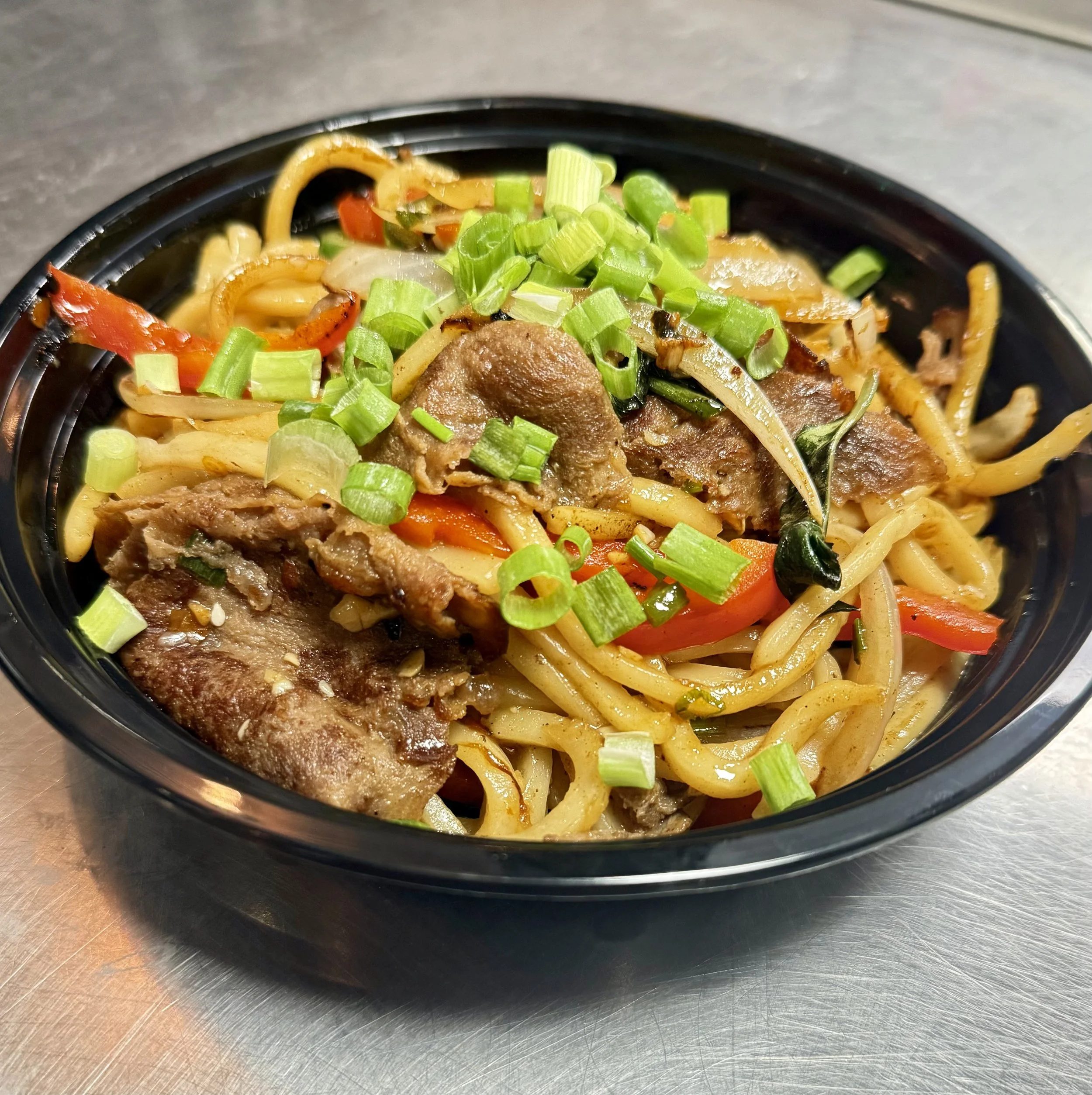 A bowl of stir-fried noodles with beef, sliced vegetables, and chopped green onions.