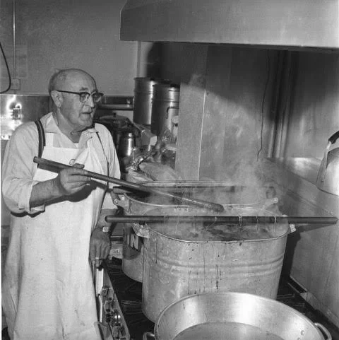 A man cooking on a stove in a kitchen, wearing glasses, an apron, and holding a spatula