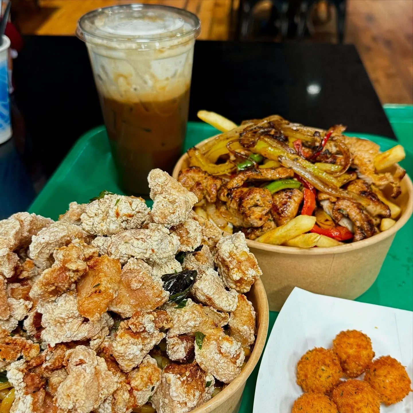 Fried chicken bites, French fries with grilled chicken and vegetables, fried croquettes, and iced coffee on a green tray.