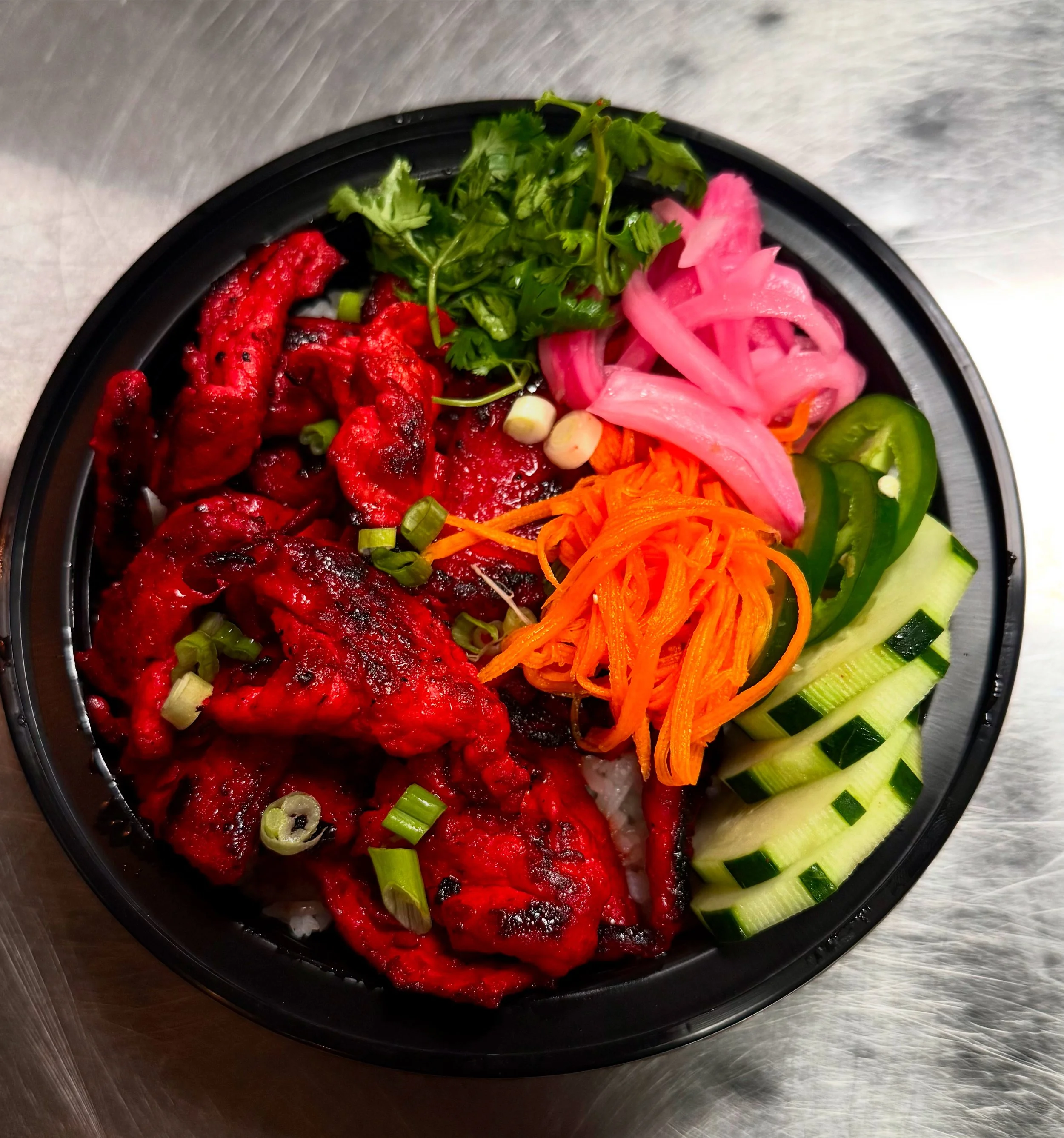 A black bowl filled with rice topped with grilled spicy chicken, garnished with green onions, along with chopped cilantro, pickled pink onions, julienned carrots, sliced green bell peppers, and sliced cucumbers.