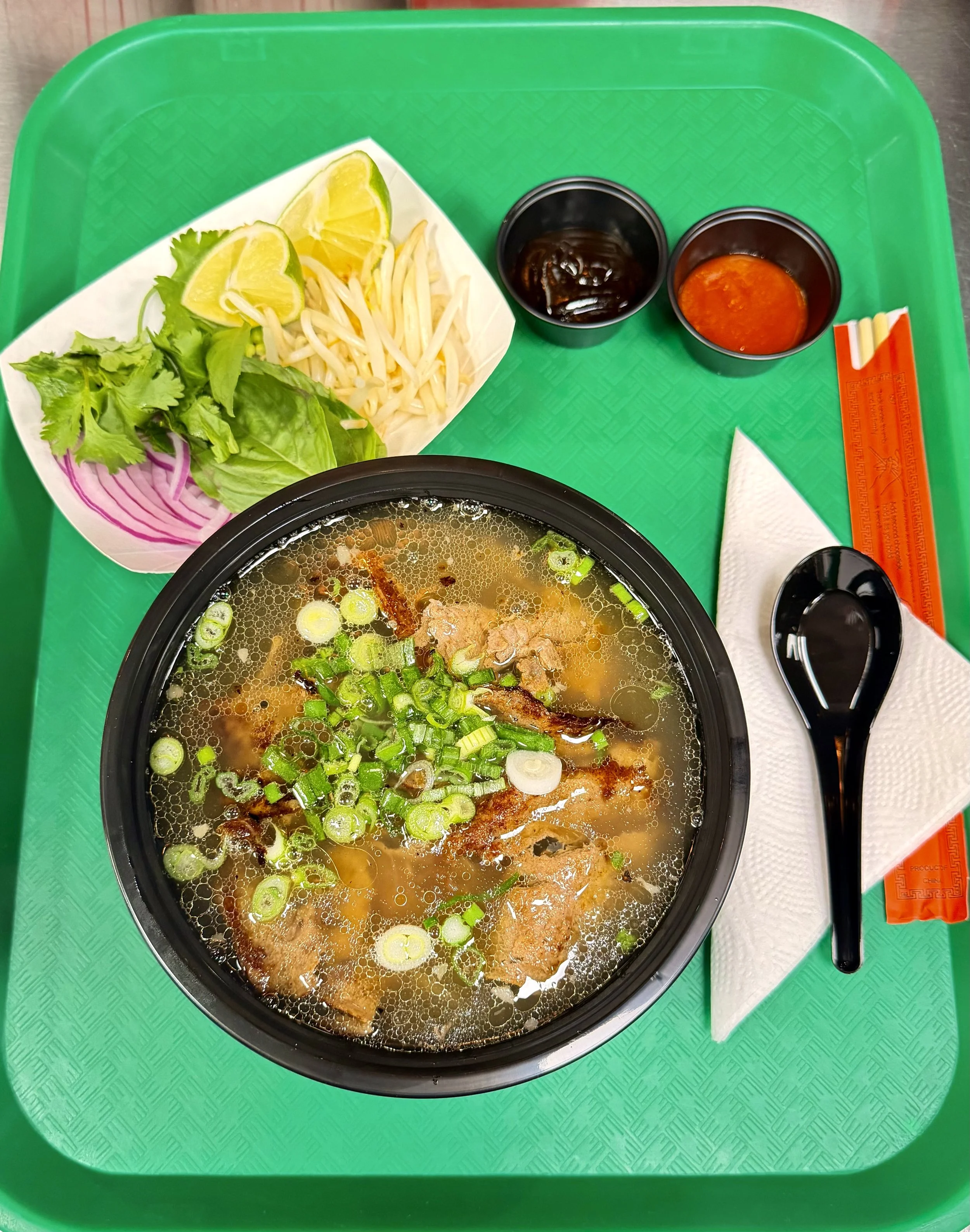A green tray with a bowl of steaming hot  topped with chopped green onions, side of lemon wedges, bean sprouts, lettuce, and sliced onions in a small dish, along with soy sauce and hot sauce in small cups, pair of orange chopsticks, and a black spoon
