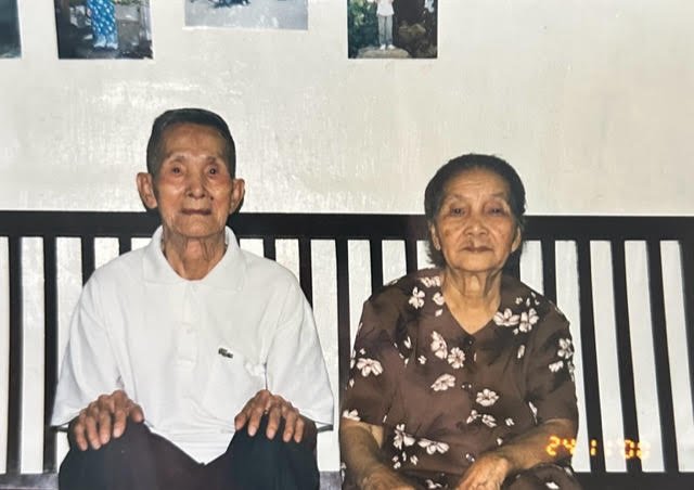 An elderly man and woman sitting on a black bench in front of a white wall with photographs. The man is wearing a white shirt, and the woman is wearing a brown floral dress.