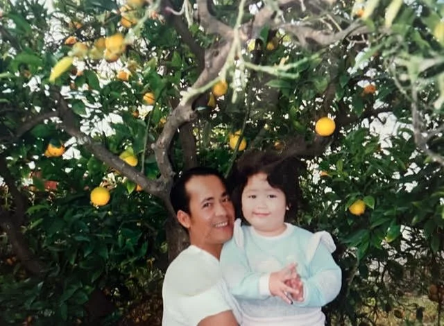 A man and a young girl standing close together in front of a lemon tree with yellow lemons, smiling at the camera.