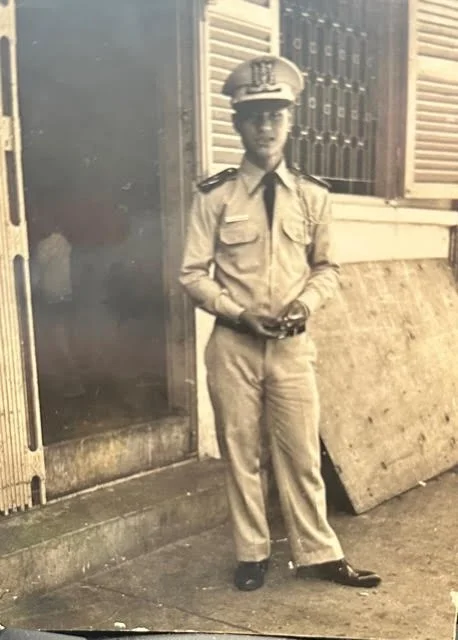 A young man in a military or police uniform standing outdoors next to a building with open shutters. He is holding an object in his hands and looking at the camera.