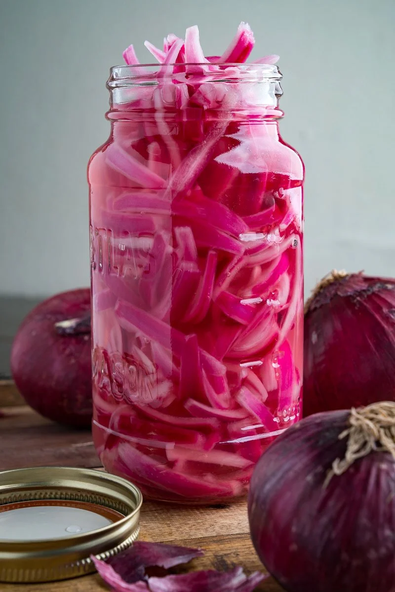 A glass jar filled with pickled red onions, with whole red onions surrounding it on a wooden surface.