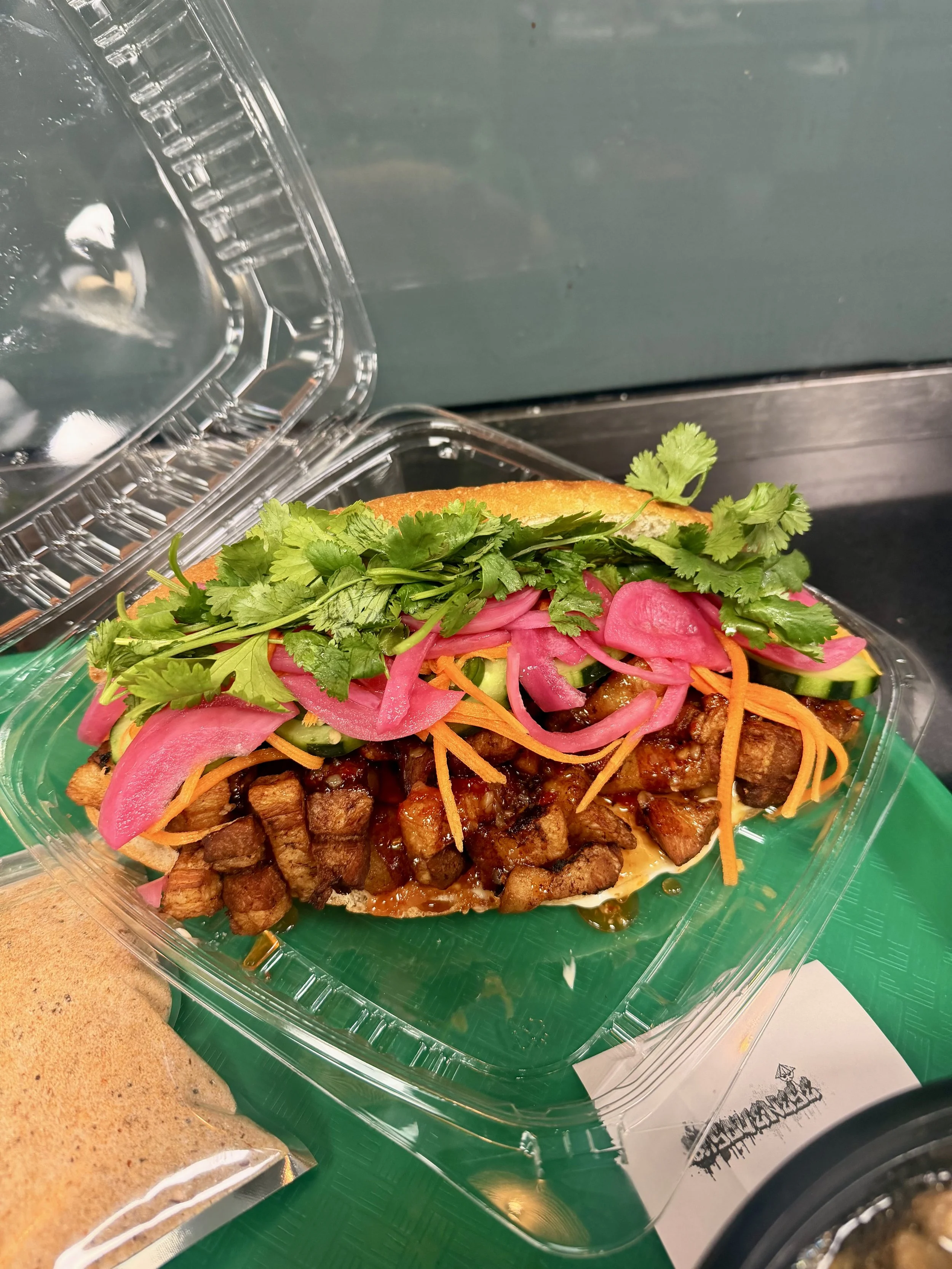 Crispy pork belly banh mi filled with  fresh cilantro, pickled pink onions, shredded carrots, and sliced cucumbers, served in a clear plastic container.