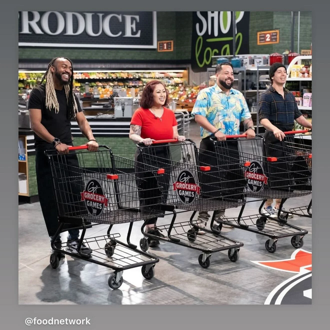 Four people with shopping carts in a grocery store, smiling and laughing as they shop. The background shows grocery shelves and signs reading 'Produce' and 'Shop Local'.