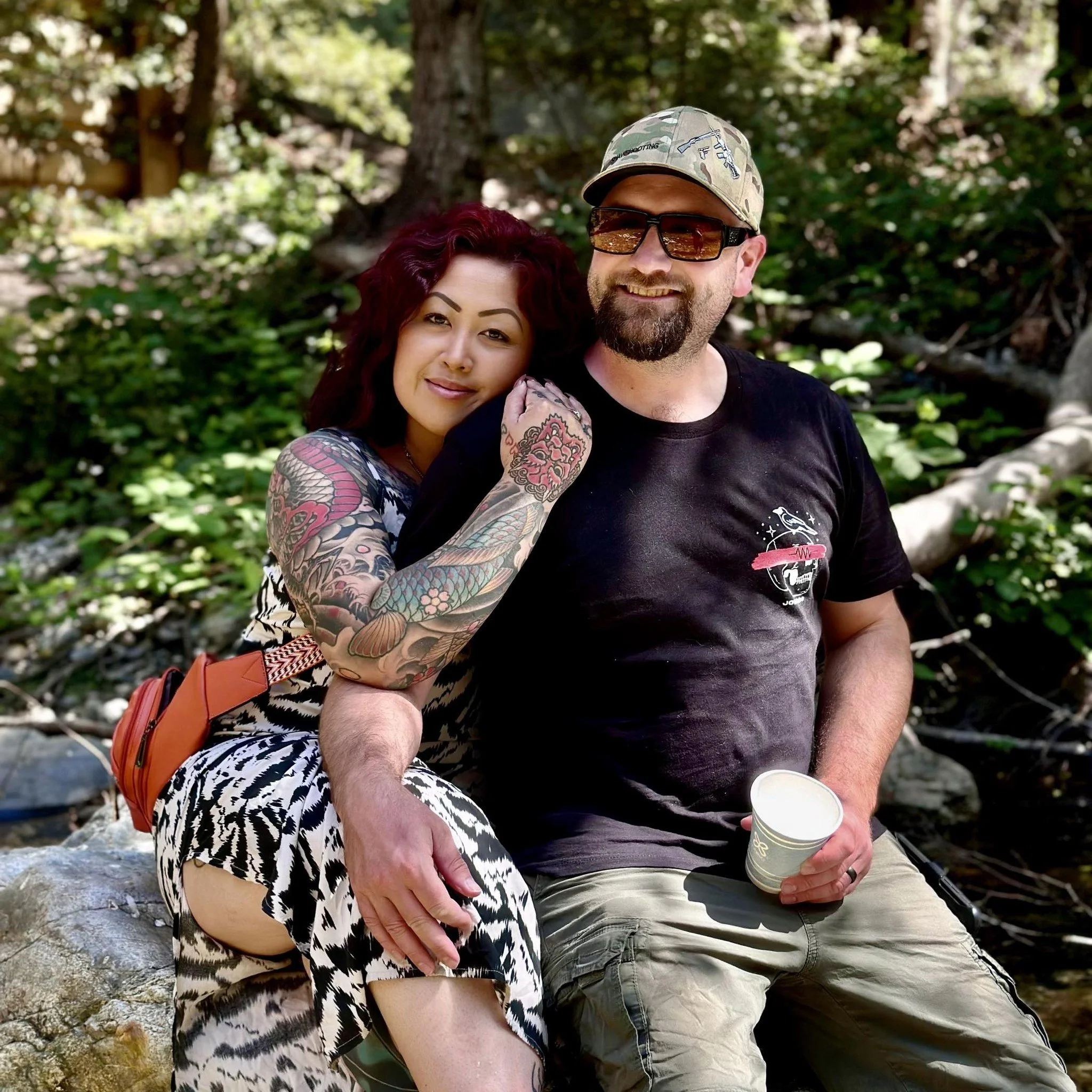 A tattooed woman with red hair and a man with a beard and sunglasses, both smiling, sitting outdoors on rocks with green foliage in the background.