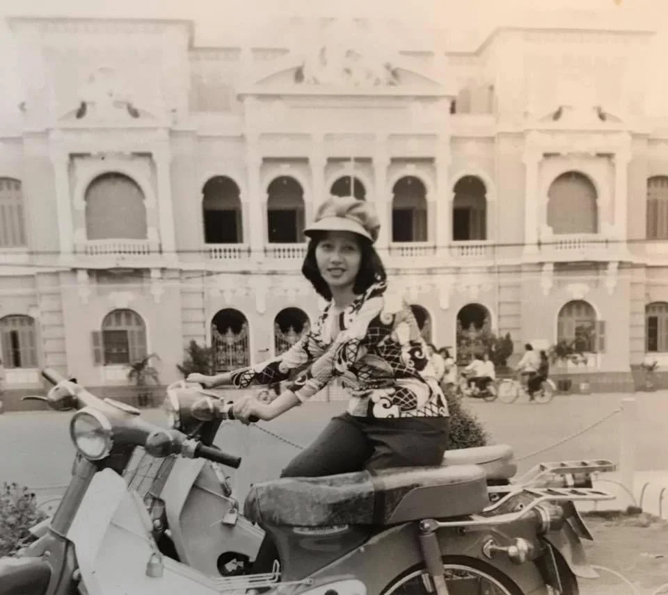 A woman sitting on a scooter in front of a large building with arches and balconies, in an urban setting with people and bicycles in the background.