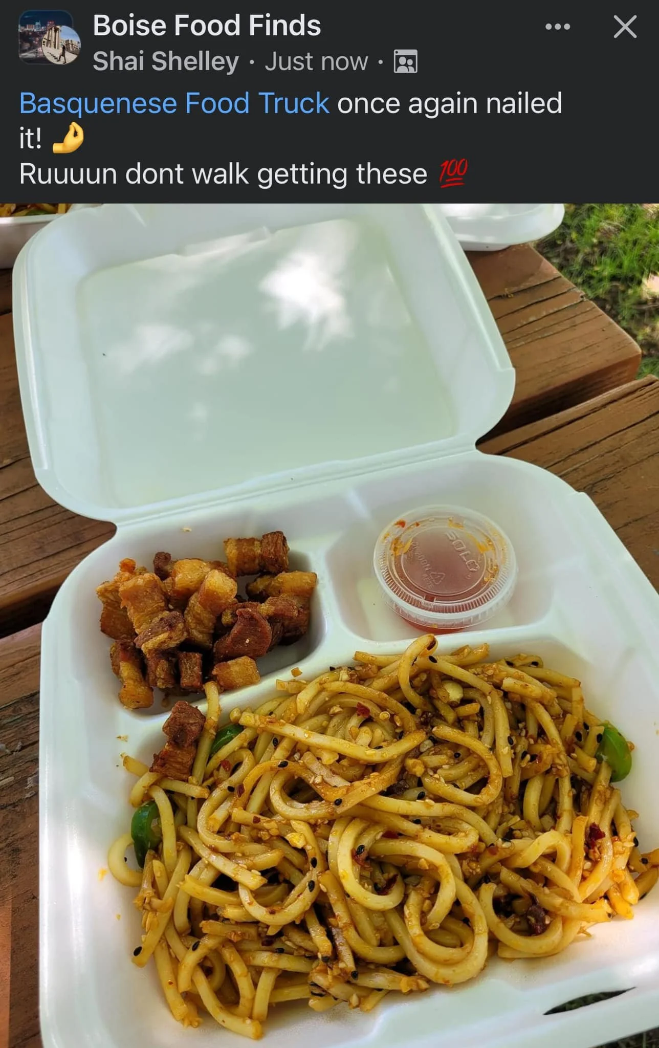 Open white takeout container with Chinese food, including fried pork belly, stir-fried noodles with vegetables, and a small container of sauce on a wooden table.