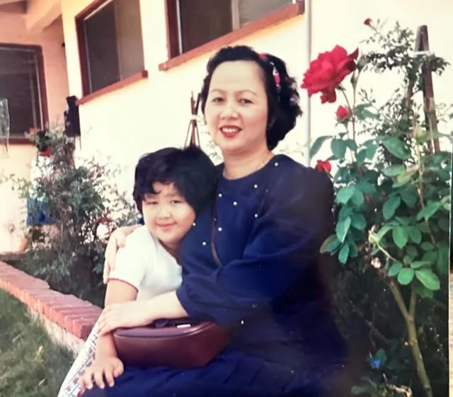 A woman and a young girl sitting outdoors near a rose bush, smiling at the camera.