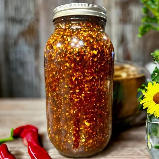 A glass jar filled with homemade chili garlic crunch condiment on a wooden surface with red chili peppers and a plant nearby.