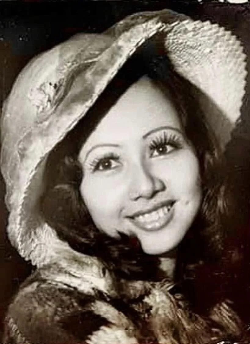 Black and white photo of a woman wearing a wide-brimmed hat with a decorative band, smiling with visible teeth, looking slightly to her left.