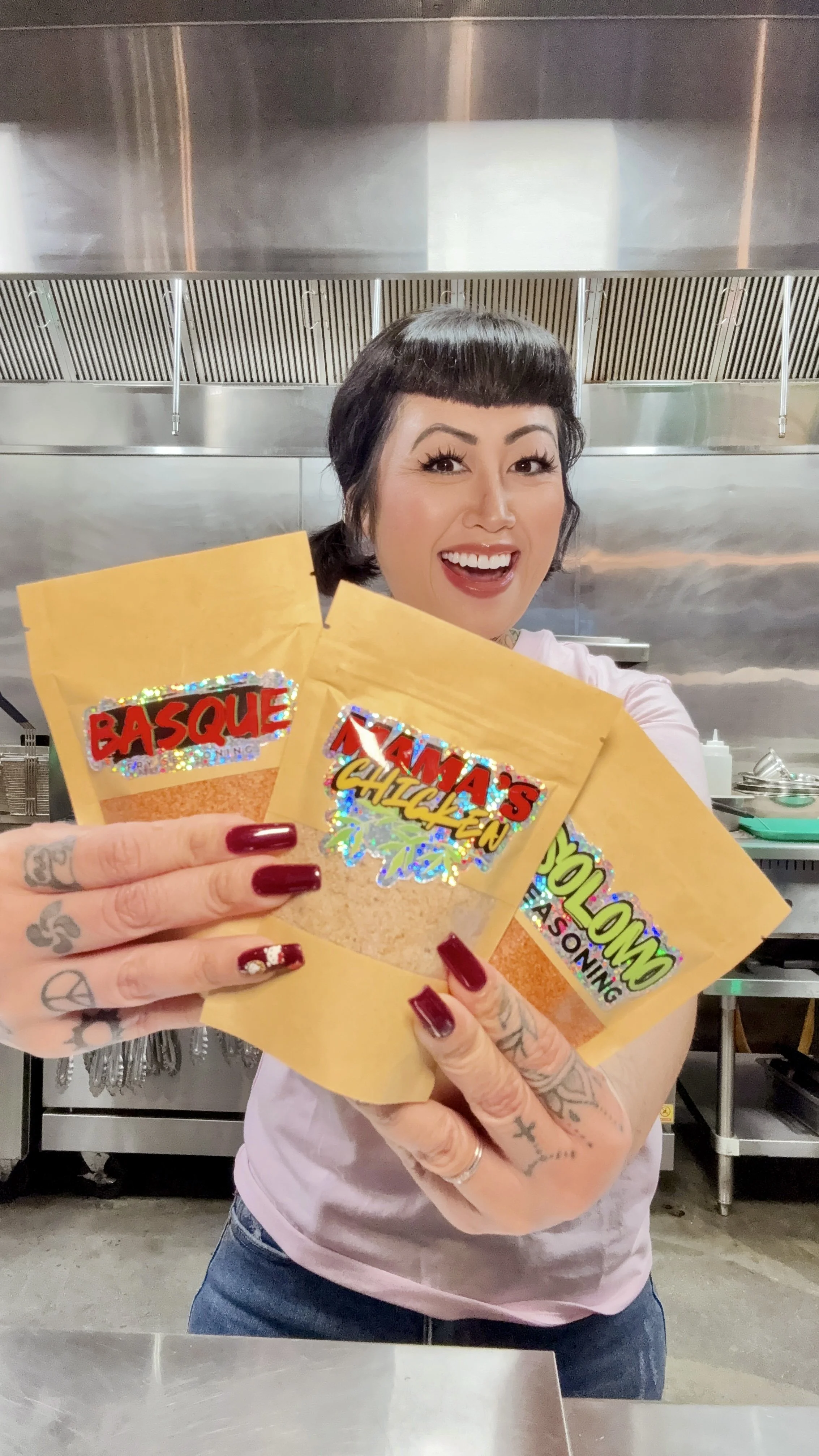 A woman with tattoos and red nails smiling and holding three spice packets labeled 'Basque', 'Lama's Chicken', and 'Cajun.' She is in a professional kitchen with stainless steel appliances in the background.